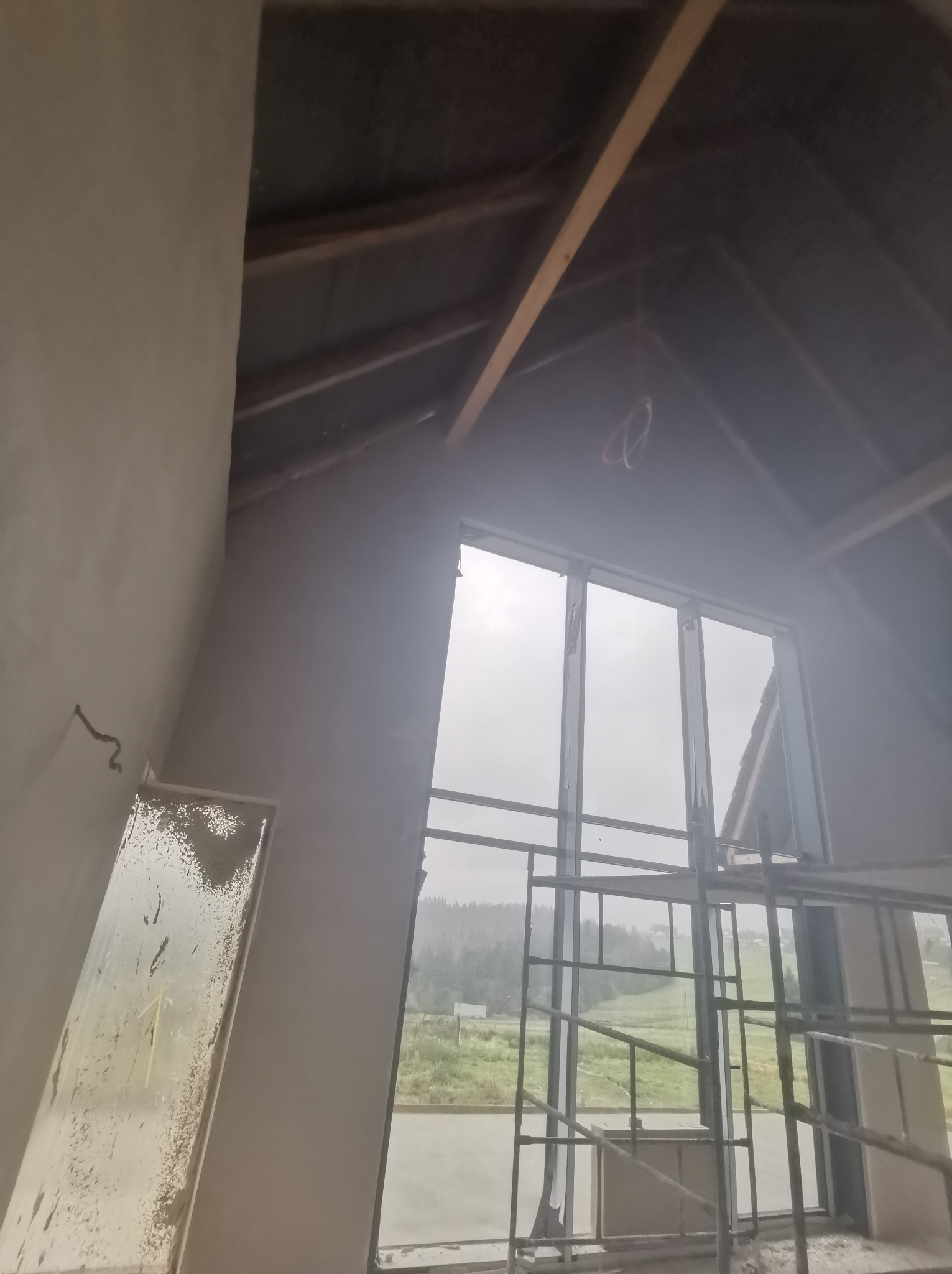 Wnętrze domu po nałożeniu szarego tynku maszynowego, widoczne okno z rusztowaniem i fragment dachu. Tynk świeży, widać zacieki. Na zewnątrz zieleń.