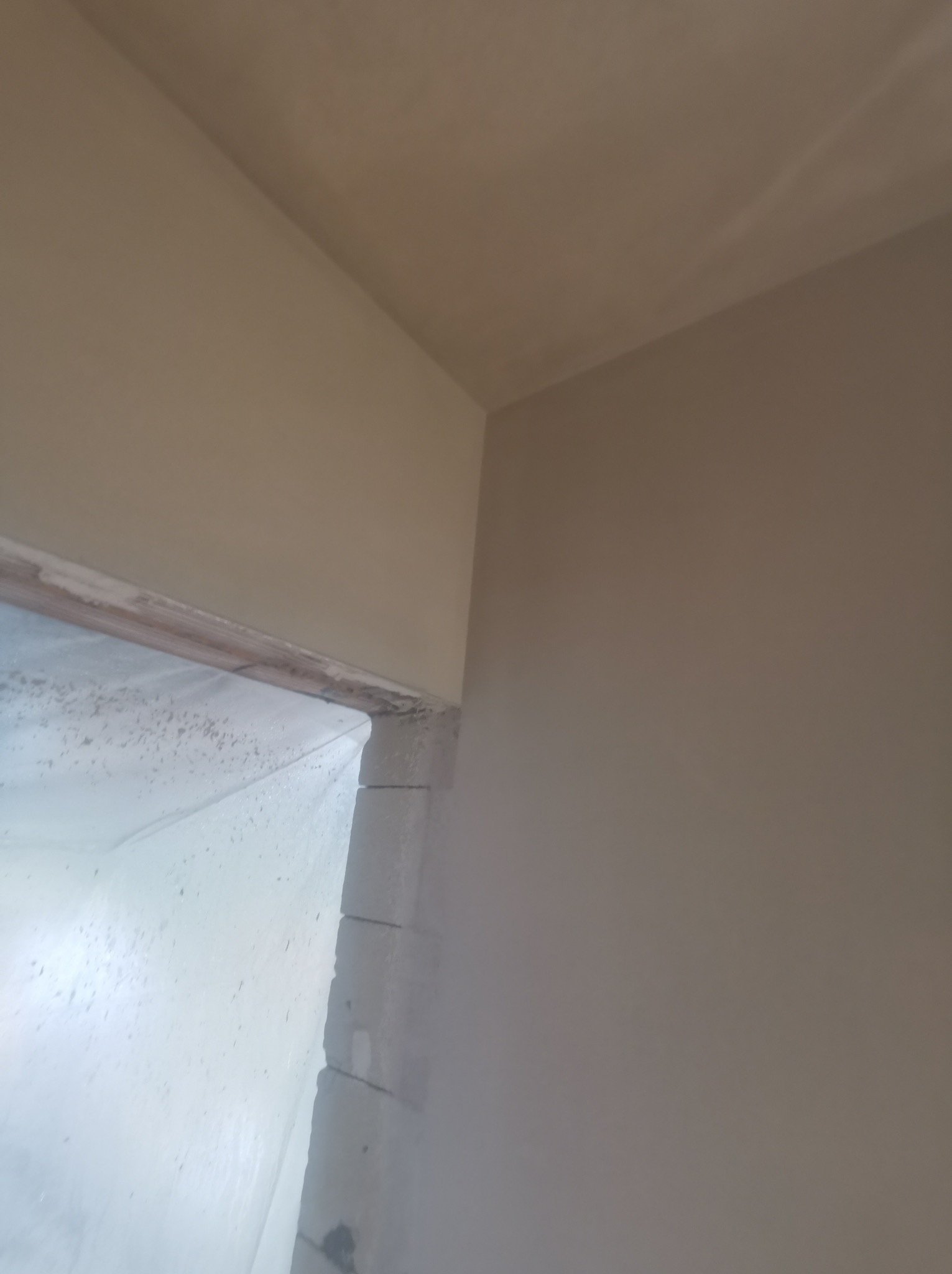 Ściana po nałożeniu gładzi gipsowej, widoczne zabezpieczone folią okno. Jasne, neutralne kolory. Precyzyjne wykończenie narożnika.