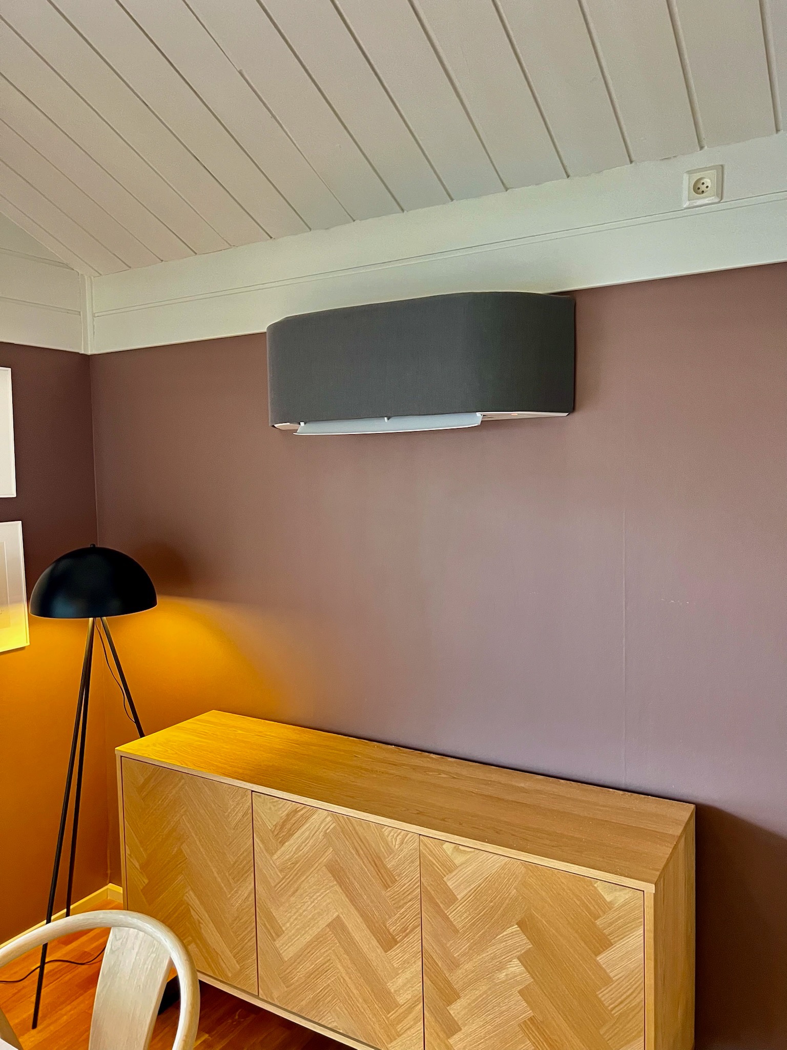Szary klimatyzator ścienny w nowoczesnym wnętrzu z komodą w jodełkę, lampą i drewnianym sufitem. Widoczny gładki, pomalowany na fioletowo fragment ściany.