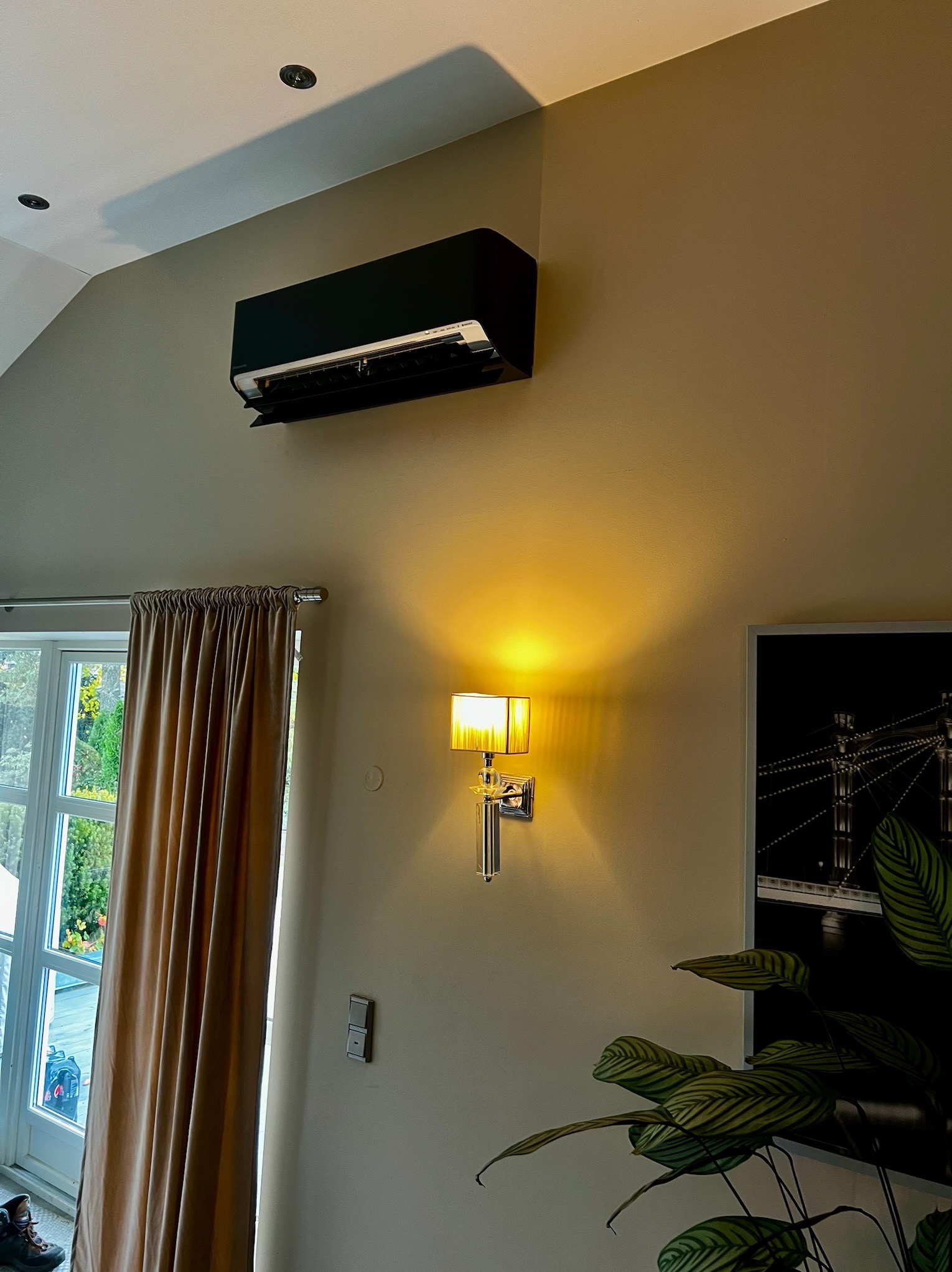 Czarny klimatyzator ścienny w jasnym wnętrzu, z lampą i obrazem w tle. Widok na okno z zasłoną i butami w rogu. Minimalistyczny design.