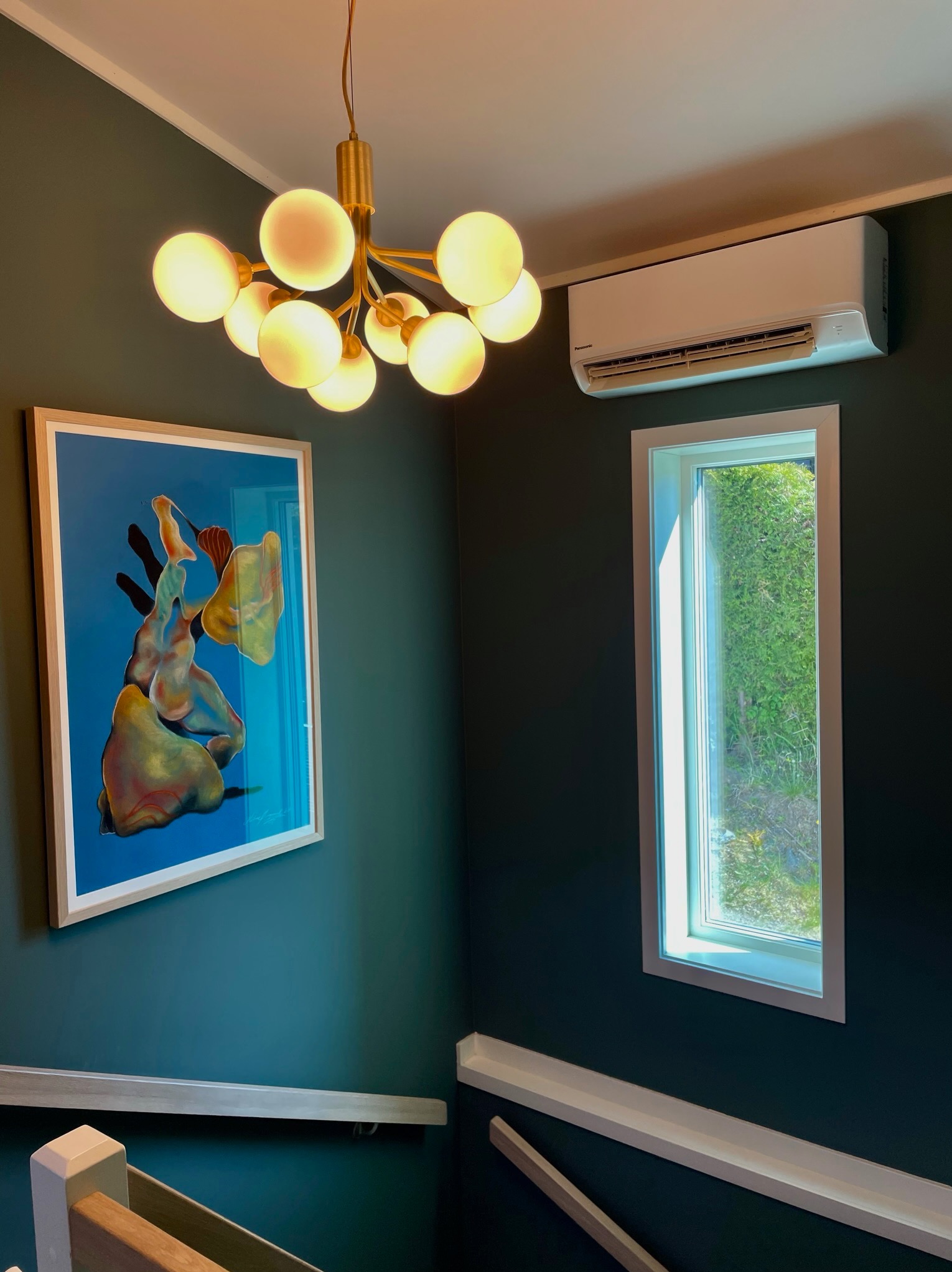 Nowoczesny klimatyzator ścienny Panasonic w jasnym wnętrzu, obok okna i obrazu, oświetlony designerską lampą. Widok z dołu schodów.