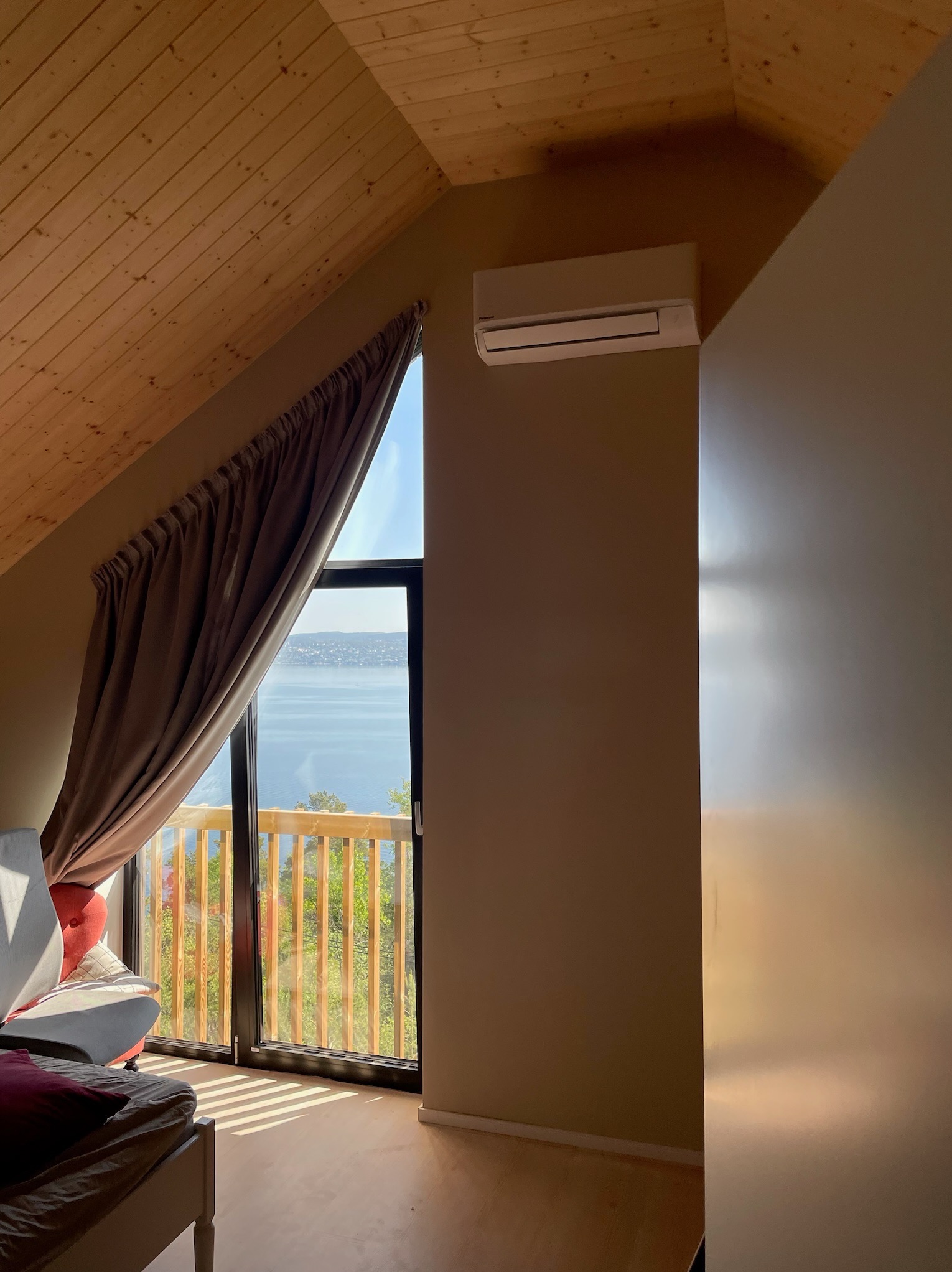 Klimatyzator ścienny w jasnym pokoju na poddaszu z drewnianym sufitem, widok na jezioro przez okno balkonowe z zasłoną.