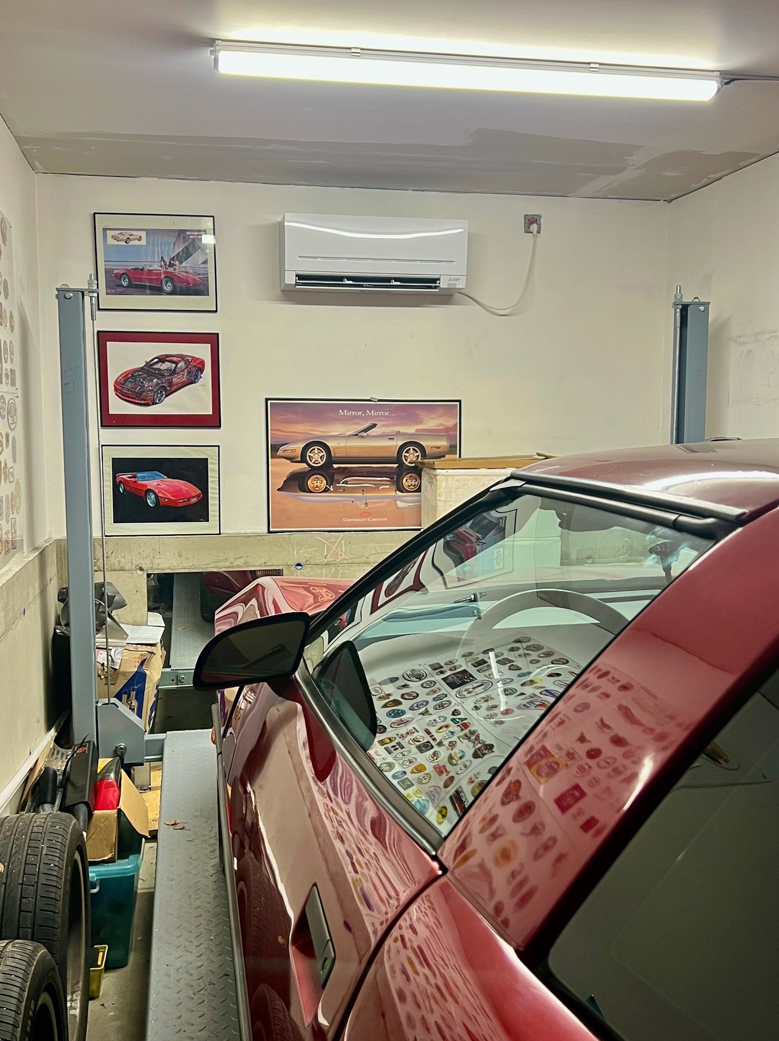 Biała klimatyzacja zamontowana w garażu nad plakatami samochodów w Wieliczce. Czerwony samochód na podnośniku garażowym, oświetlenie LED.
