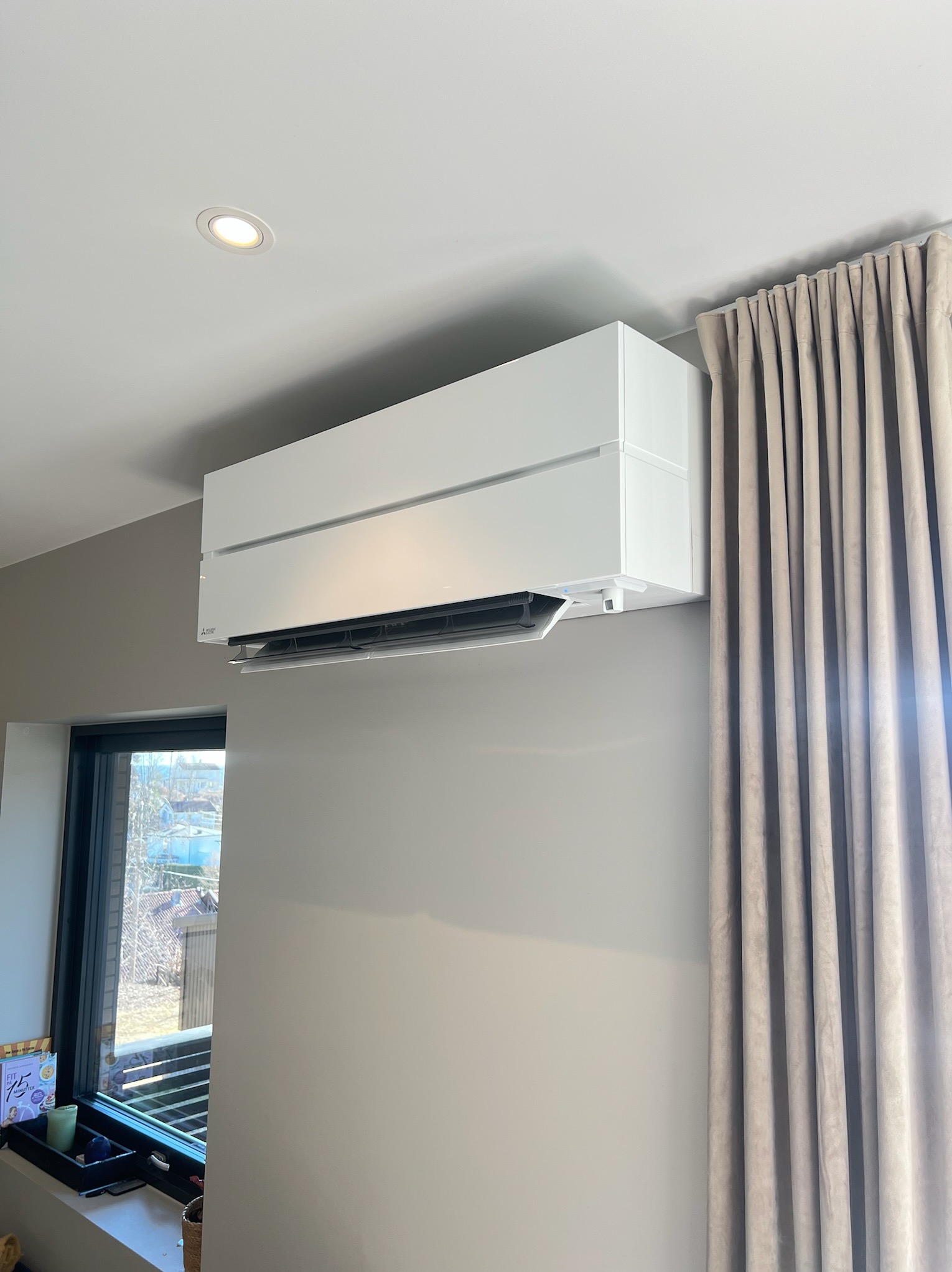 Nowoczesny, biały klimatyzator ścienny Mitsubishi w jasnym wnętrzu, obok okna i zasłon. Widoczny element dekoracyjny na parapecie.