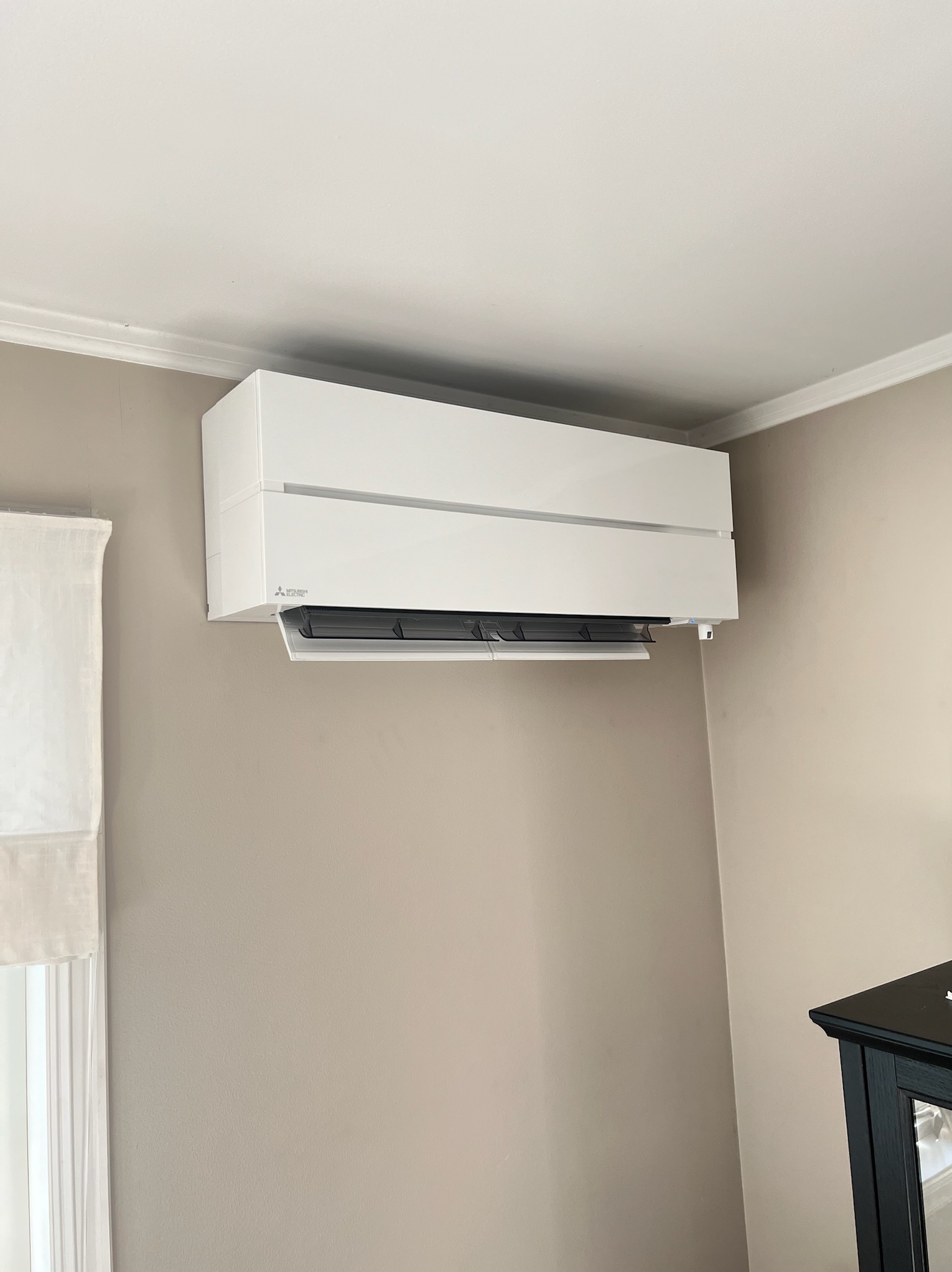 Biały klimatyzator Mitsubishi Electric zamontowany na beżowej ścianie w jasnym wnętrzu. Widoczny fragment okna z zasłoną i czarny mebel.