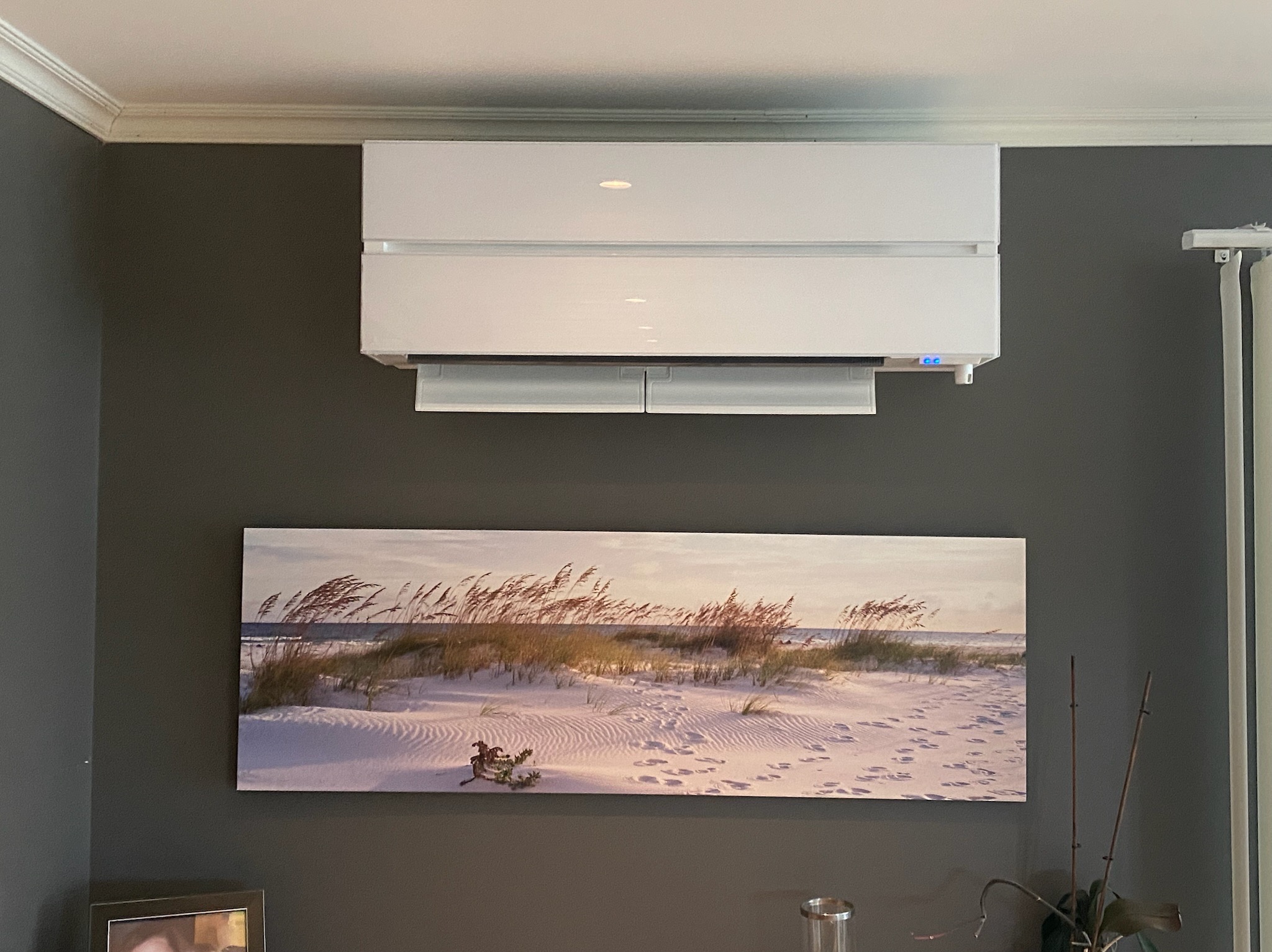 Biały klimatyzator ścienny z delikatnym podświetleniem, umieszczony nad obrazem plaży w pokoju o szarych ścianach.