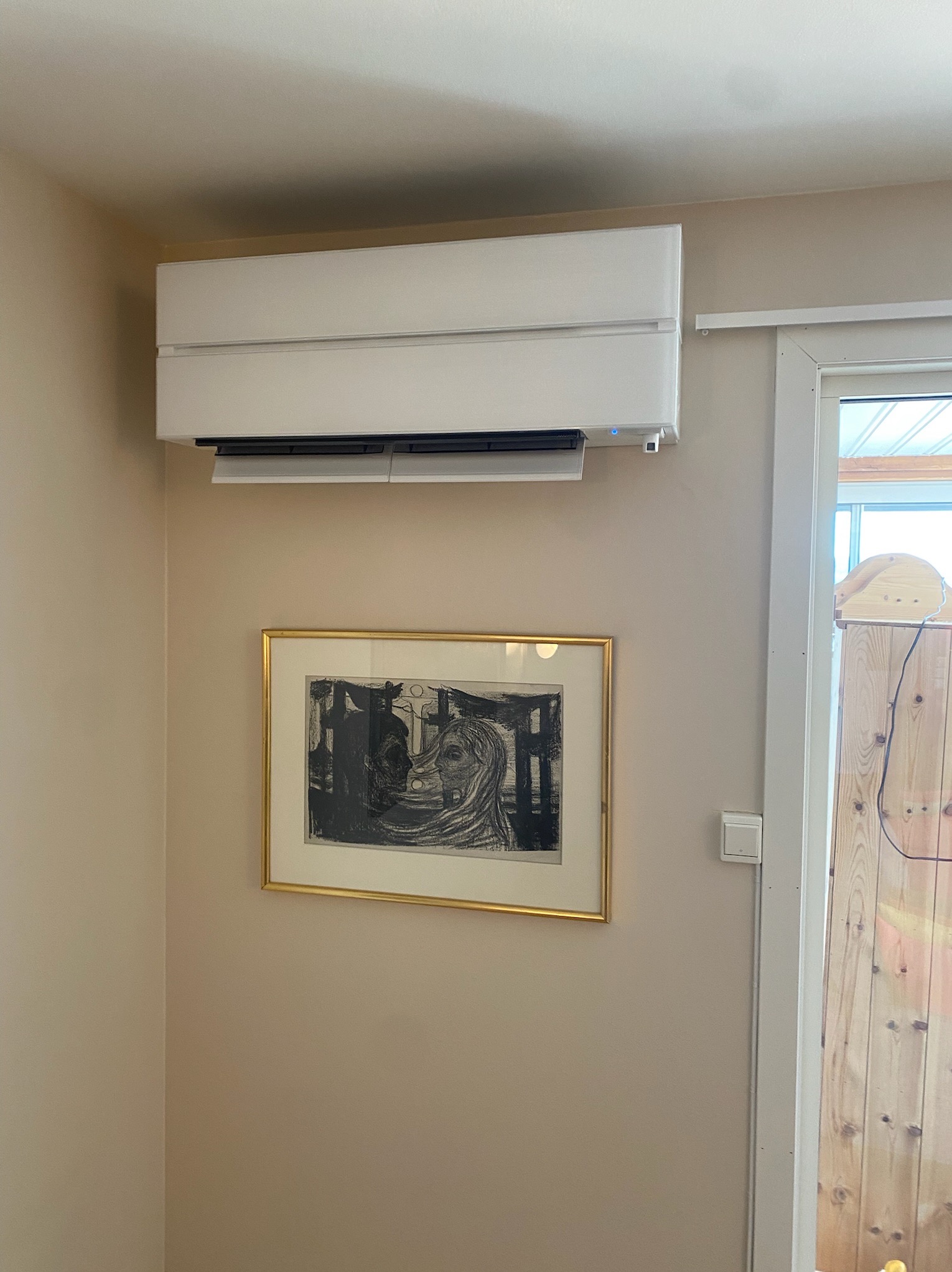 Biały klimatyzator ścienny zamontowany nad obrazem w złotej ramie na beżowej ścianie w domu. Widoczny fragment okna i drewnianych drzwi.