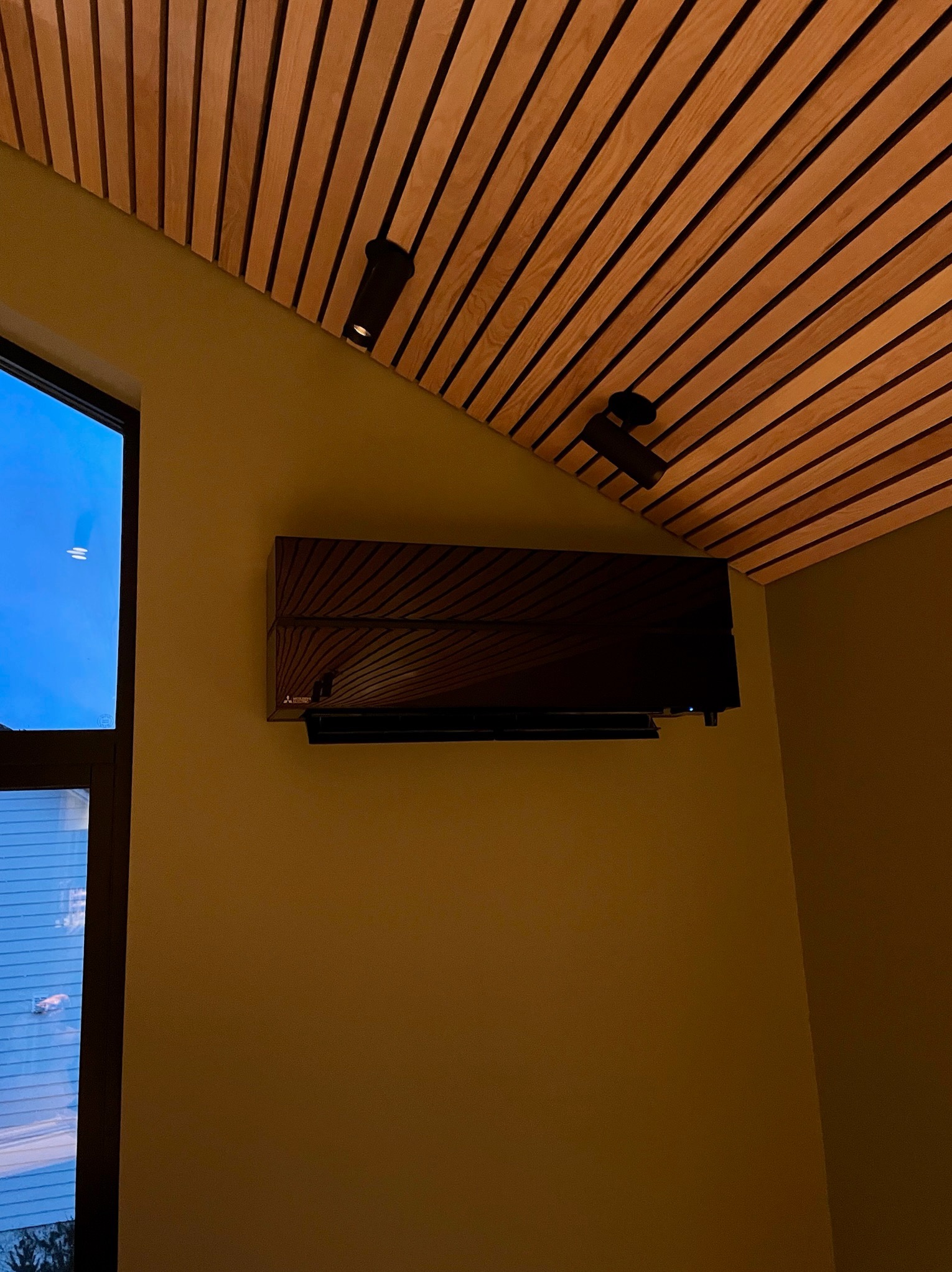 Czarny klimatyzator ścienny Mitsubishi w nowoczesnym wnętrzu z drewnianym sufitem i oknem z widokiem na zewnątrz. Minimalistyczny design i oświetlenie punktowe.