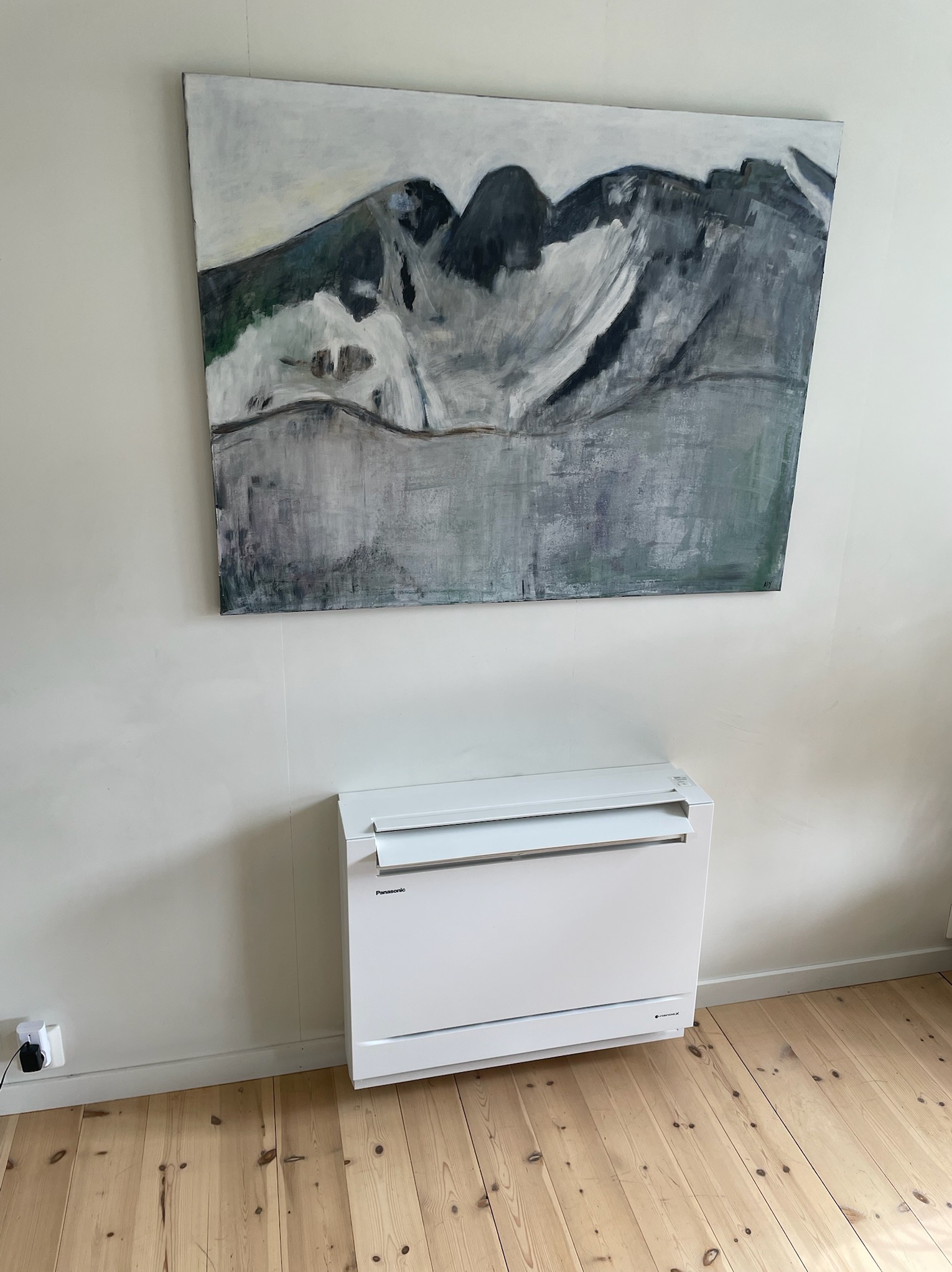 Nowoczesny, biały klimatyzator podłogowy Panasonic na tle ściany z obrazem górskim, na drewnianej podłodze. Widoczny kabel zasilający.