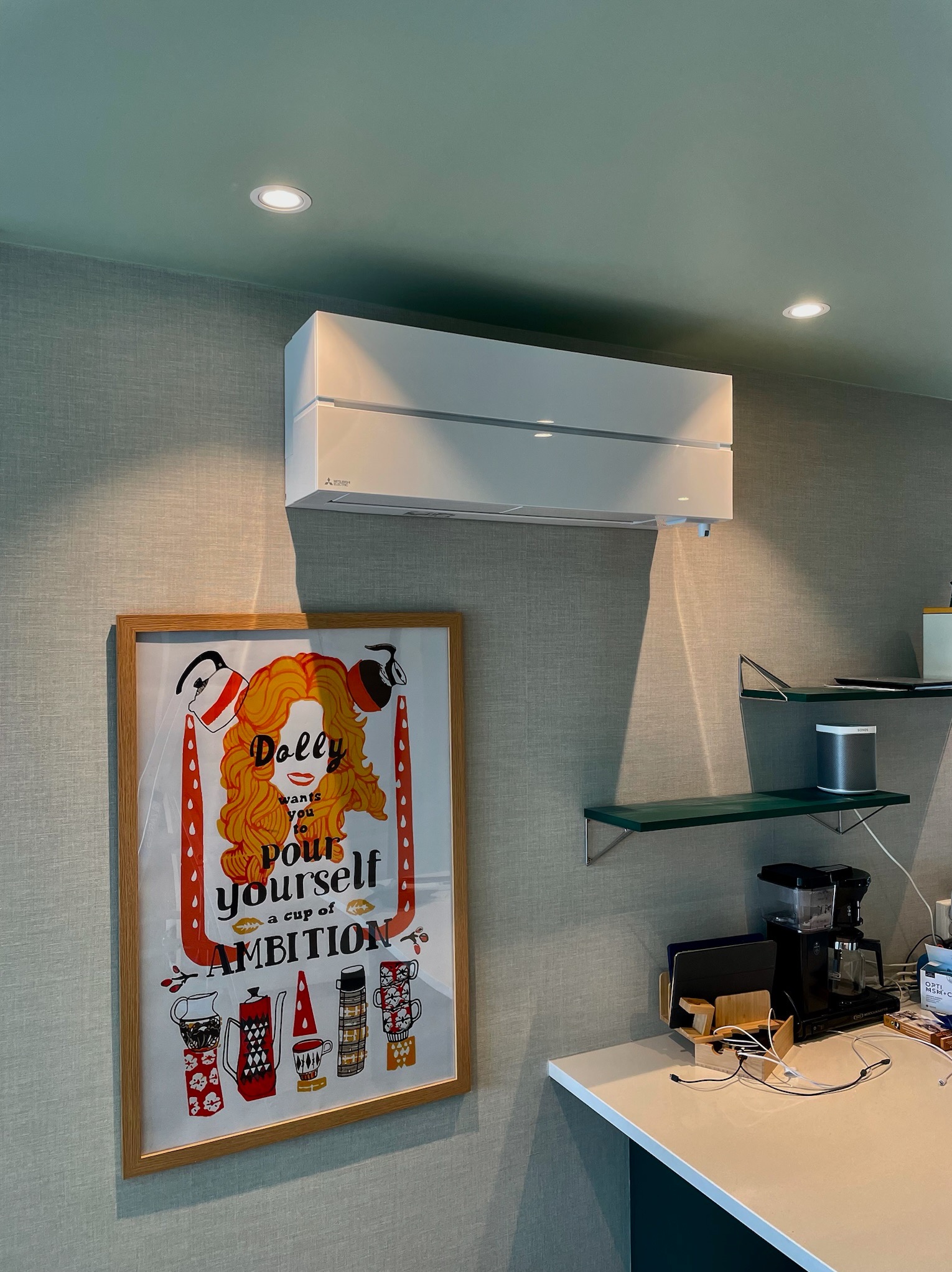 Nowoczesny, biały klimatyzator Mitsubishi zamontowany na ścianie nad oprawionym plakatem w pokoju z zielonymi akcentami i półkami.