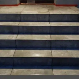 Wykończone schody zewnętrzne z szarych, kwadratowych płytek. Widoczne fugi i detale wykończenia przy wejściu. Ujęcie z dołu, podkreślające regularność wzoru.