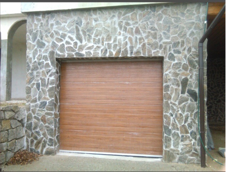 Brama garażowa z okładziną kamienną z nieregularnych elementów. Zastosowano brązową bramę segmentową. Widoczny fragment elewacji budynku.
