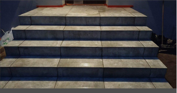 Wykończone schody zewnętrzne z szarych, kwadratowych płytek. Widoczne fugi i detale wykończenia przy wejściu. Ujęcie z dołu, podkreślające regularność wzoru.