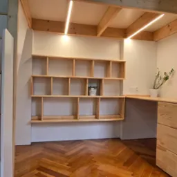 Funkcjonalna zabudowa z surowego drewna: biurko z szufladami, półki na ścianie, oświetlenie LED i terrarium w ramie, parkiet w jodełkę. Widok zza rogu.