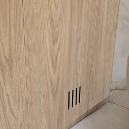 Arkadia - Fragment szafki z jasnego drewna z czterema pionowymi szczelinami wentylacyjnymi u dołu, widoczna podłoga i fragment ściany z płytek.