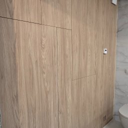 Arkadia - Drewniana zabudowa w łazience z ukrytymi drzwiami i panelem sterowania. Naturalna kolorystyka drewna, minimalistyczny design, widoczne detale wykończenia.