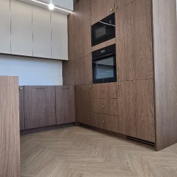 Nowoczesna kuchnia z drewnianymi szafkami, wbudowanymi piekarnikami i mikrofalówką. Parkiet w jodełkę. Minimalistyczne oświetlenie sufitowe.