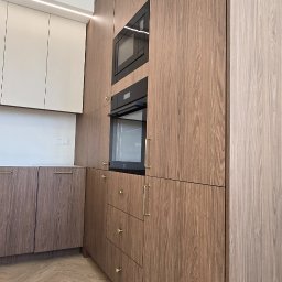 Nowoczesna kuchnia z drewnianymi frontami, złotymi uchwytami i wbudowanym sprzętem AGD. Parkiet ułożony w jodełkę. Ujęcie z perspektywy podłogi.