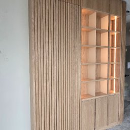 Arkadia - Nowoczesna zabudowa ścienna z jasnego drewna dębowego z pionowymi lamelami, zintegrowana z podświetlanym regałem i dolnymi szafkami. Ciepłe oświetlenie LED akcentuje półki.