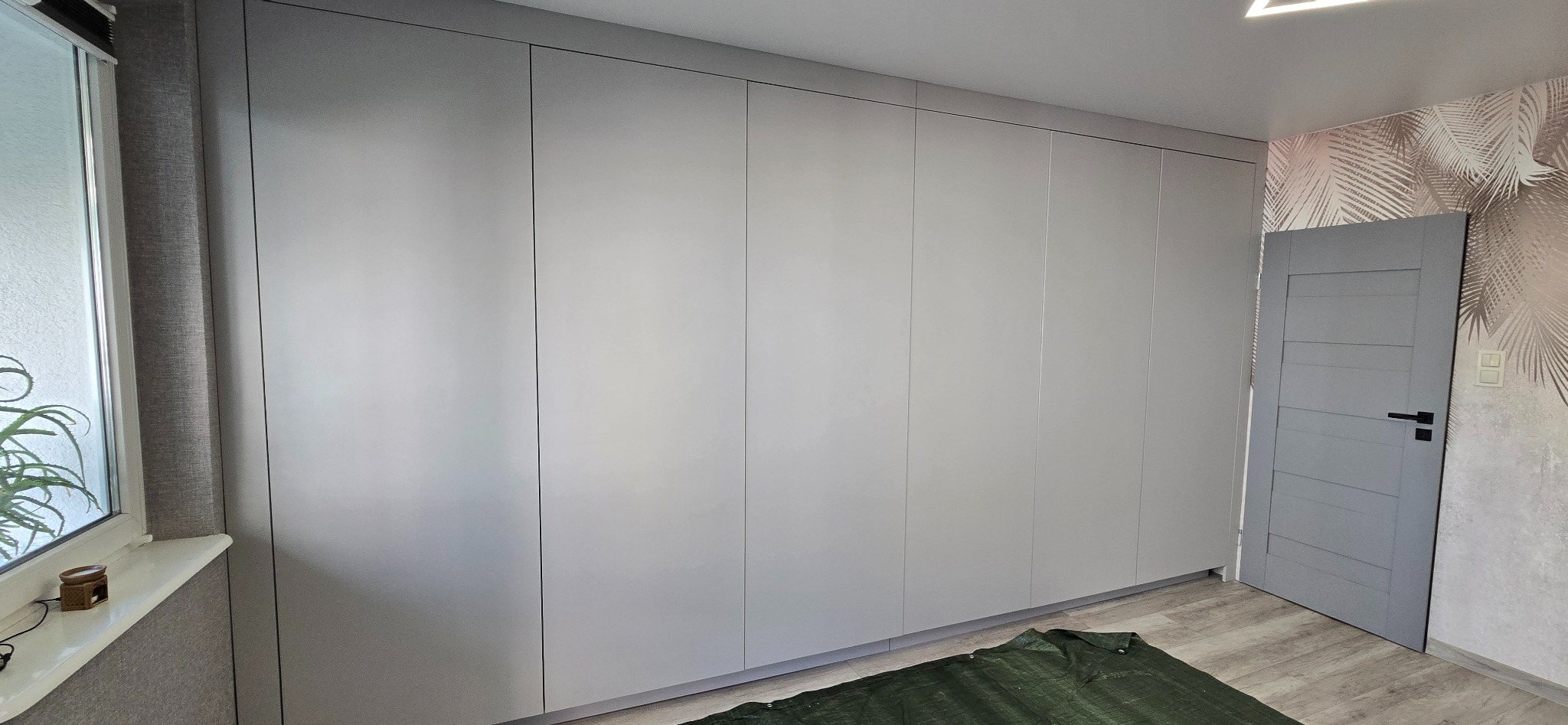 Szara szafa na wymiar z drzwiami bez uchwytów, na podłodze zielona plandeka zabezpieczająca. Widoczna szara ściana i drzwi z czarną klamką.