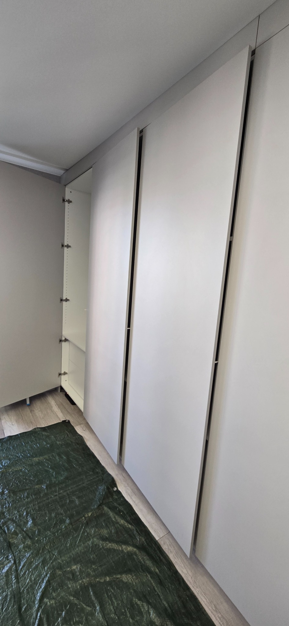 Biała szafa wnękowa z częściowo otwartymi drzwiami, zabezpieczona folią ochronną na podłodze, widok z boku. Minimalistyczny design i jasne wnętrze.