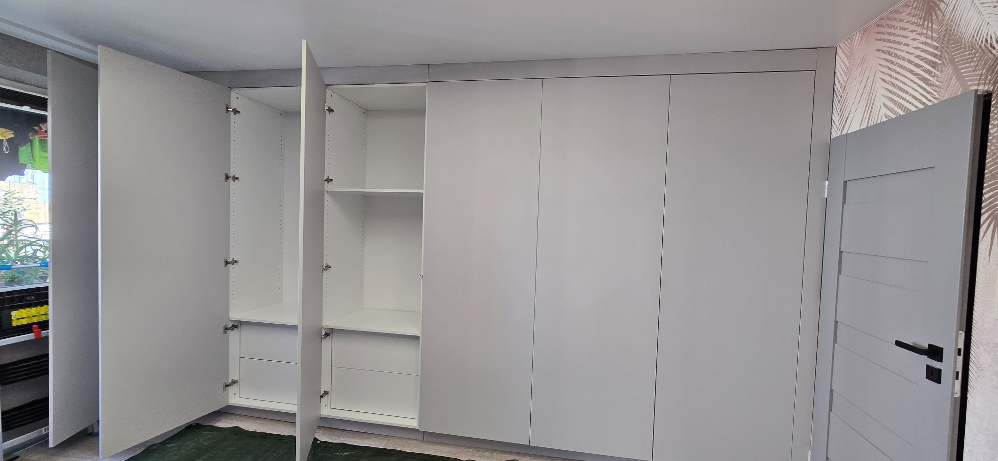 Biała szafa na wymiar z otwartymi drzwiami ukazującymi półki i szuflady, minimalistyczny design, jasne wnętrze, drzwi po prawej stronie. Tapeta w tle.