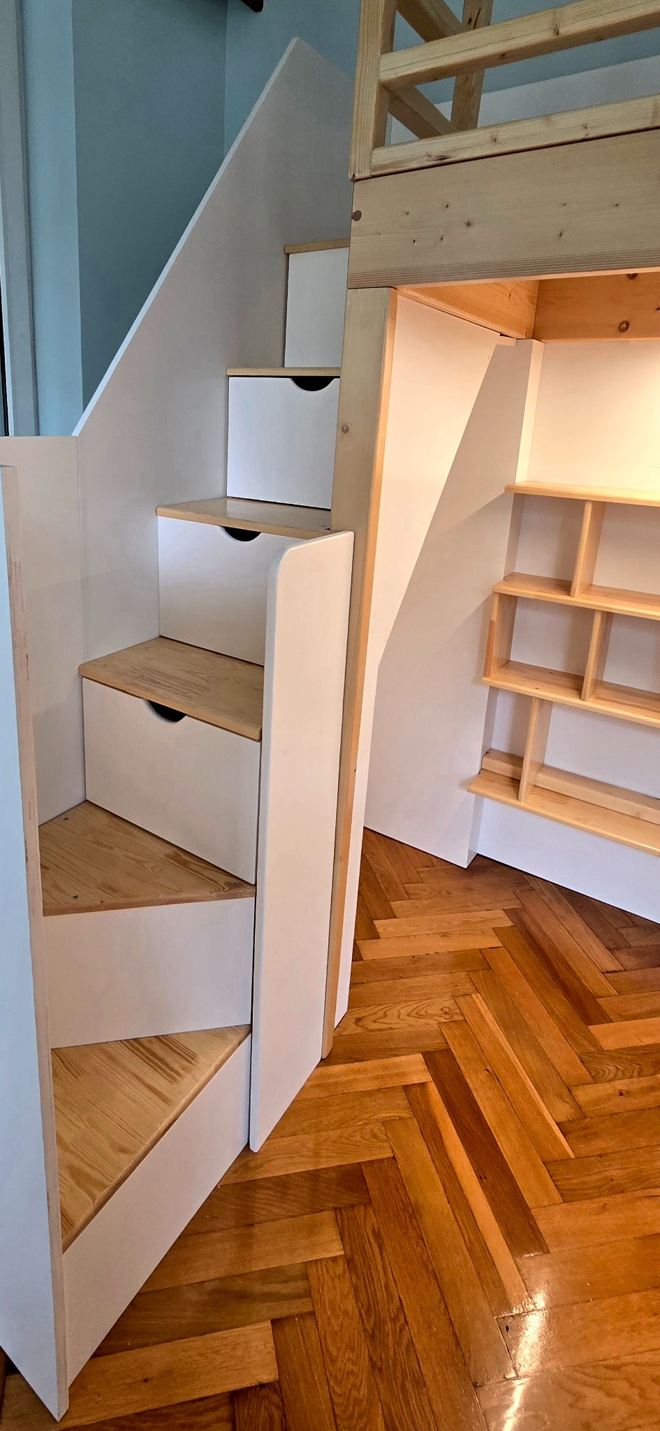 Drewniane schody z szufladami, prowadzące do antresoli, z półkami wnękowymi. Parkiet w jodełkę. Białe fronty szuflad i wykończenie. Naturalne drewno.