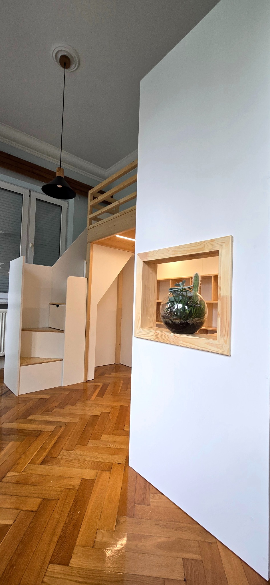 Biała antresola z drewnianymi elementami i schodami w pokoju z parkietem, z dekoracyjną wnęką na ścianie. Nowoczesne oświetlenie sufitowe.