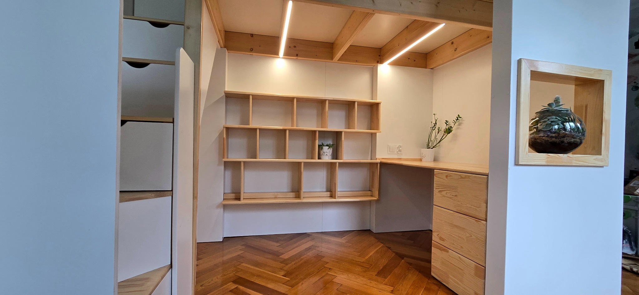 Funkcjonalna zabudowa z surowego drewna: biurko z szufladami, półki na ścianie, oświetlenie LED i terrarium w ramie, parkiet w jodełkę. Widok zza rogu.