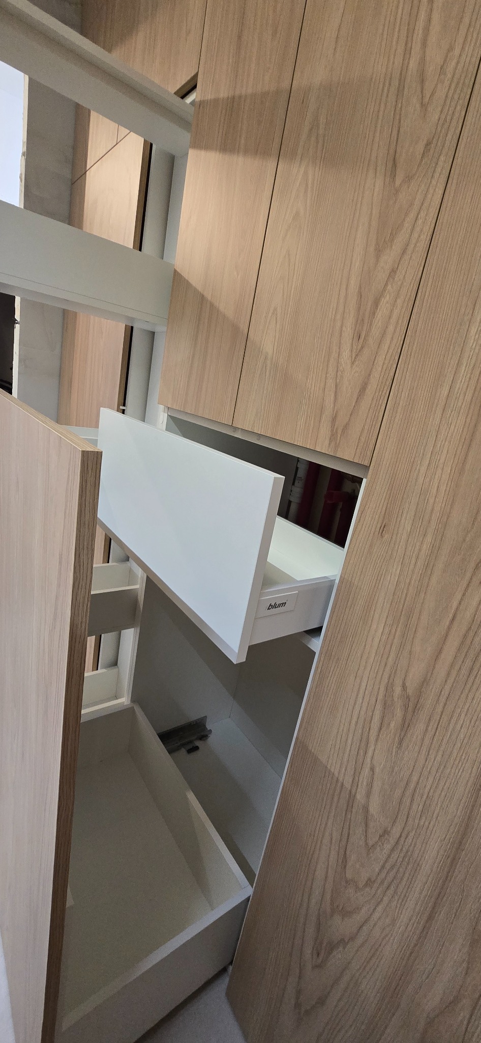 Otwarta szuflada z białym frontem w szafie z okleiną drewnopodobną. Widoczne prowadnice Blum. Ujęcie z dołu, podkreślające funkcjonalność i nowoczesny design.