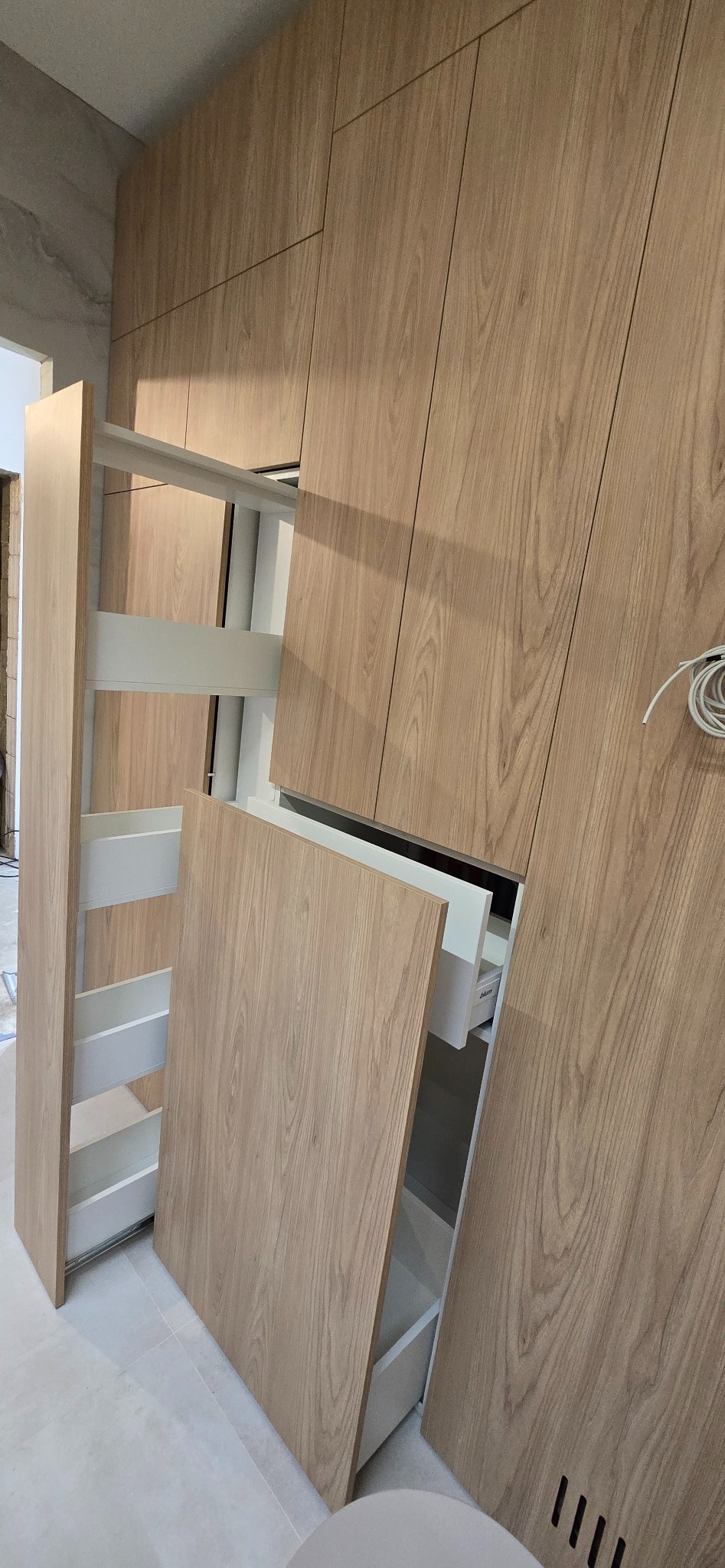 Jasna, drewniana szafa z wysuwanymi półkami i szufladami. Widoczne detale wykończenia i systemy przechowywania. Nowoczesny design, minimalistyczny styl.