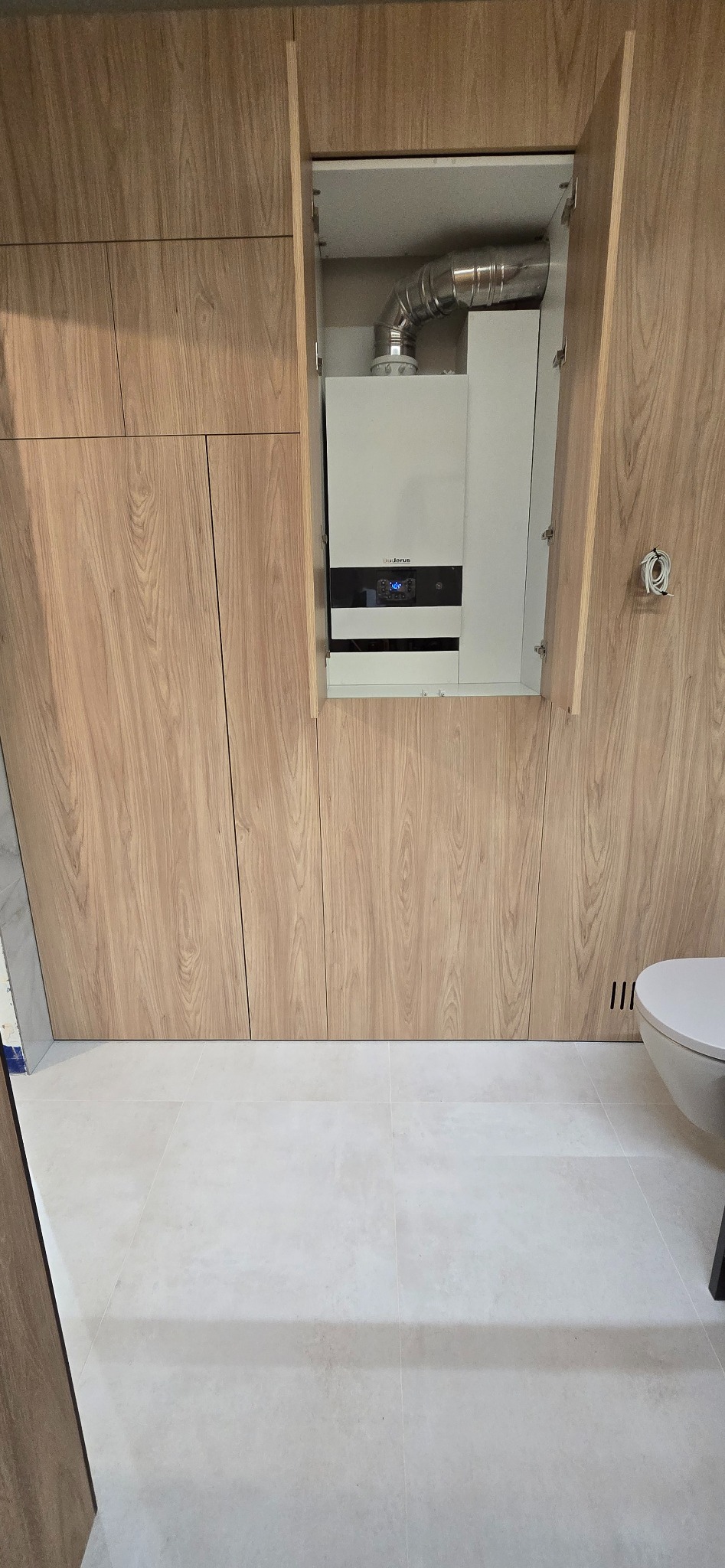 Szafa na wymiar z ukrytym piecem gazowym w łazience. Drewnopodobne fronty, białe płytki podłogowe, fragment toalety. Estetyczne rozwiązanie.