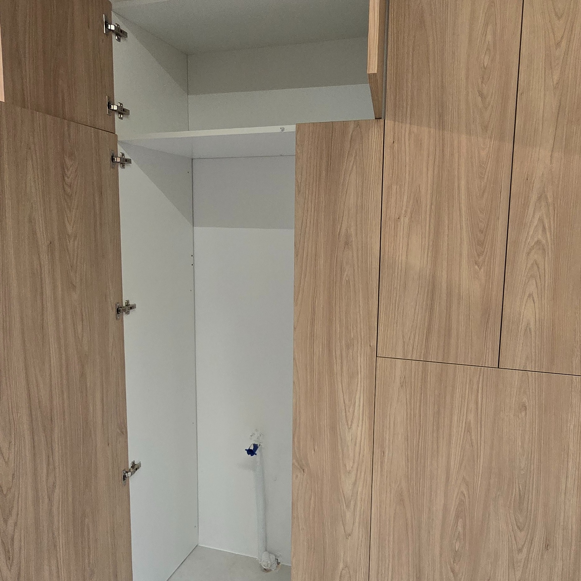 Jasna szafa z drewnianymi frontami, otwarte drzwi ukazują białe wnętrze z półką. Widoczny fragment instalacji na ścianie wewnątrz szafy.