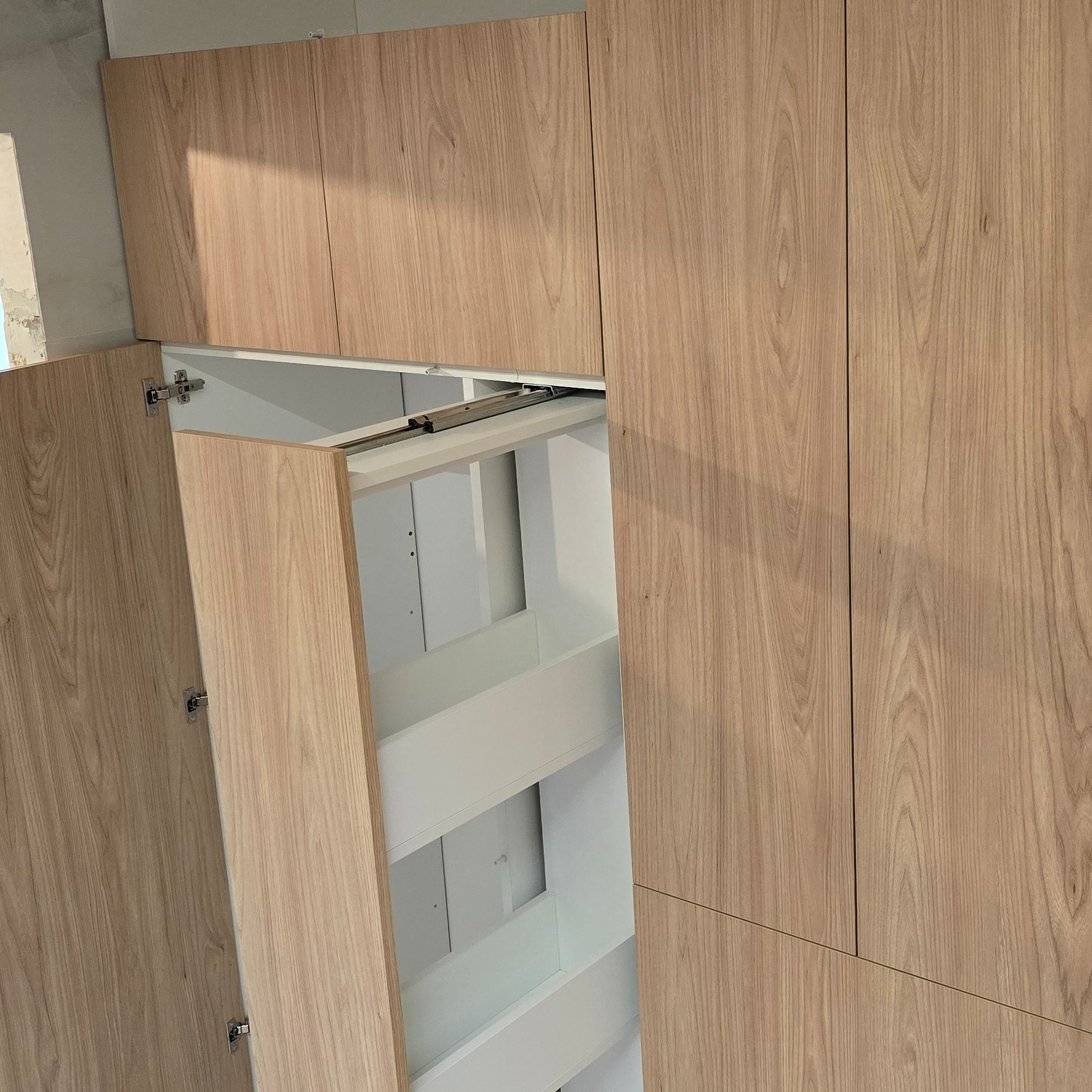 Jasna, drewnopodobna szafa z wysuwanym, białym systemem półek cargo. Widoczne zawiasy i prowadnice. Minimalistyczny design, surowy stan wykończenia wnętrza.