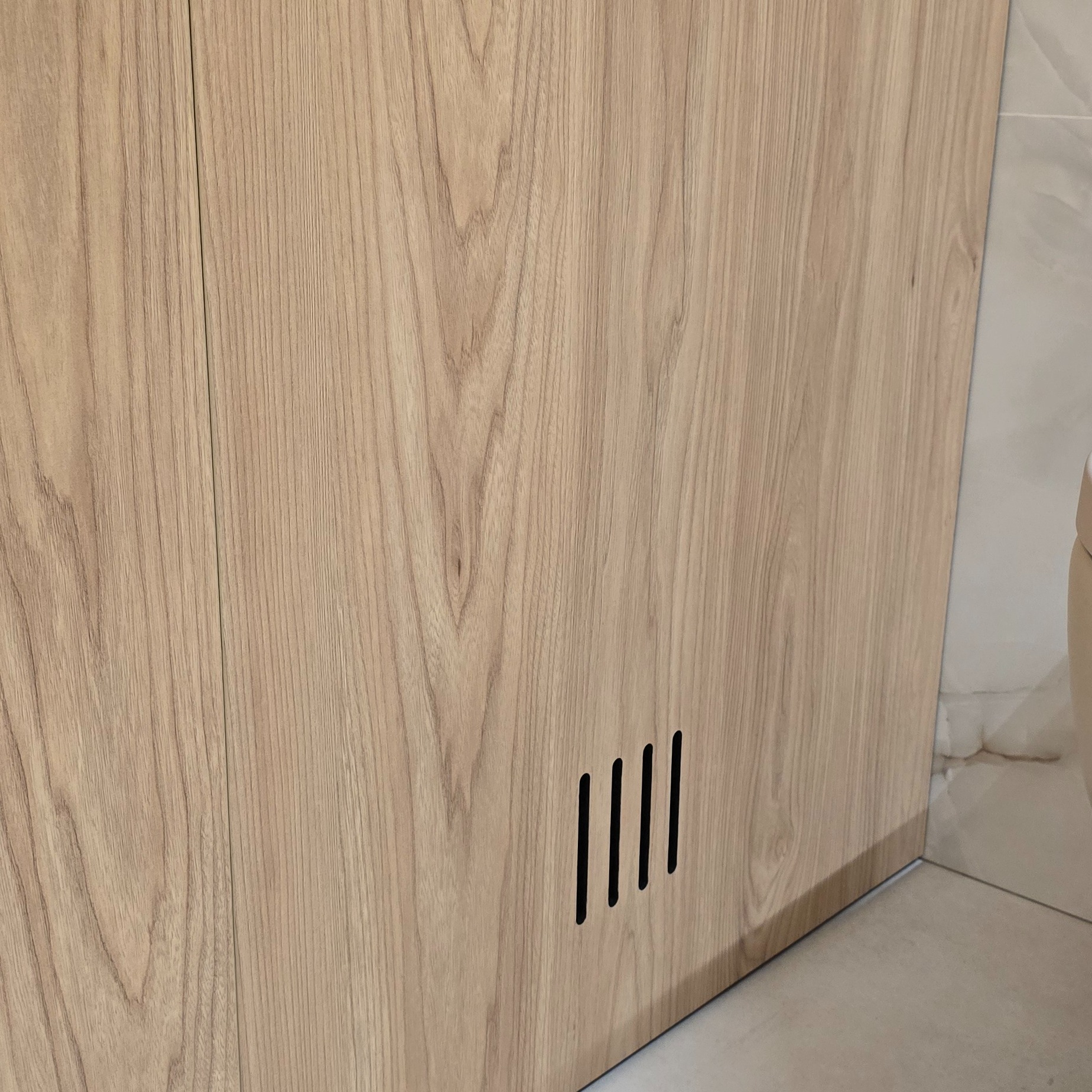 Fragment szafki z jasnego drewna z czterema pionowymi szczelinami wentylacyjnymi u dołu, widoczna podłoga i fragment ściany z płytek.