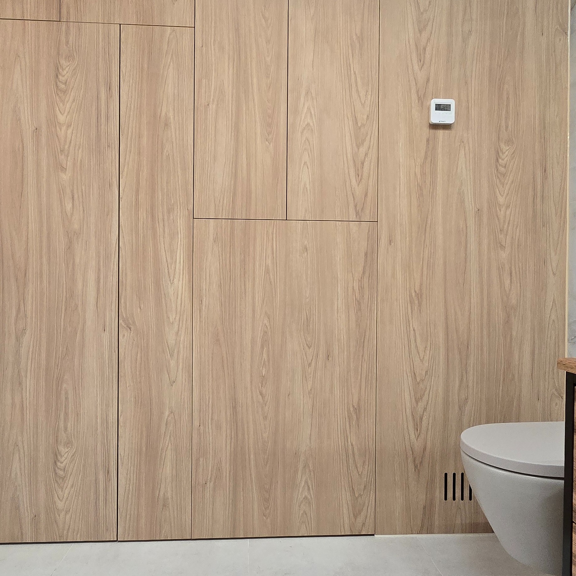 Drewniana zabudowa ściany w łazience z ukrytymi drzwiami, widoczny fragment toalety i termostat na ścianie. Minimalistyczny design, jasne drewno.