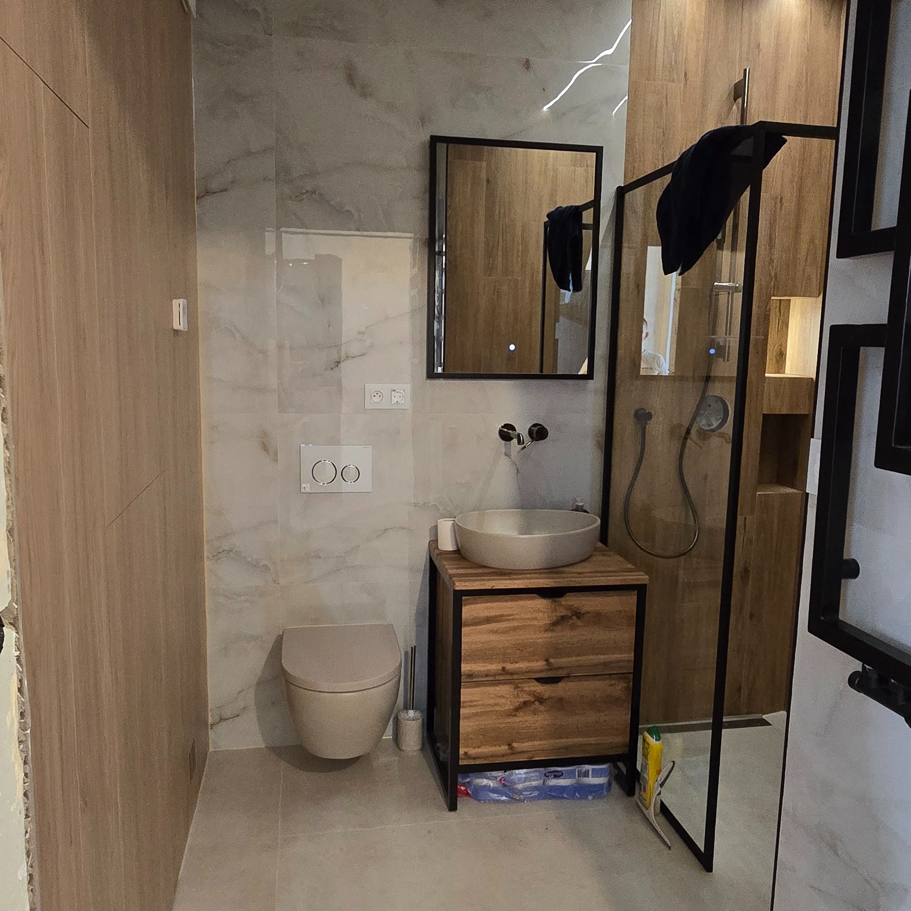 Nowoczesna łazienka z drewnianą szafką pod umywalką, wiszącą toaletą i kabiną prysznicową z czarnymi ramami. Marmurowe płytki na ścianach i podłodze.