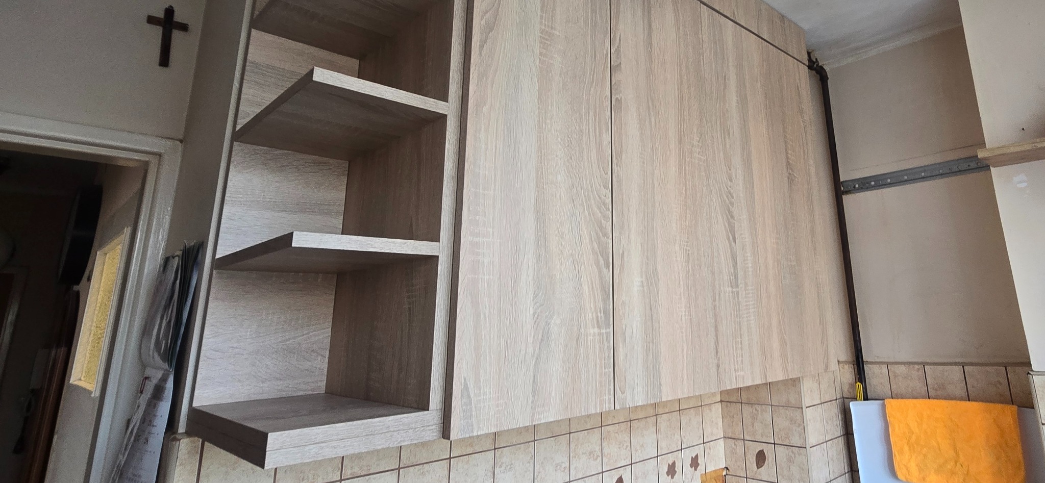 Jasne, drewnopodobne szafki kuchenne z otwartymi półkami, widoczne na tle płytek. U góry krzyż na ścianie. Ujęcie z dołu, podkreślające wysokość zabudowy.