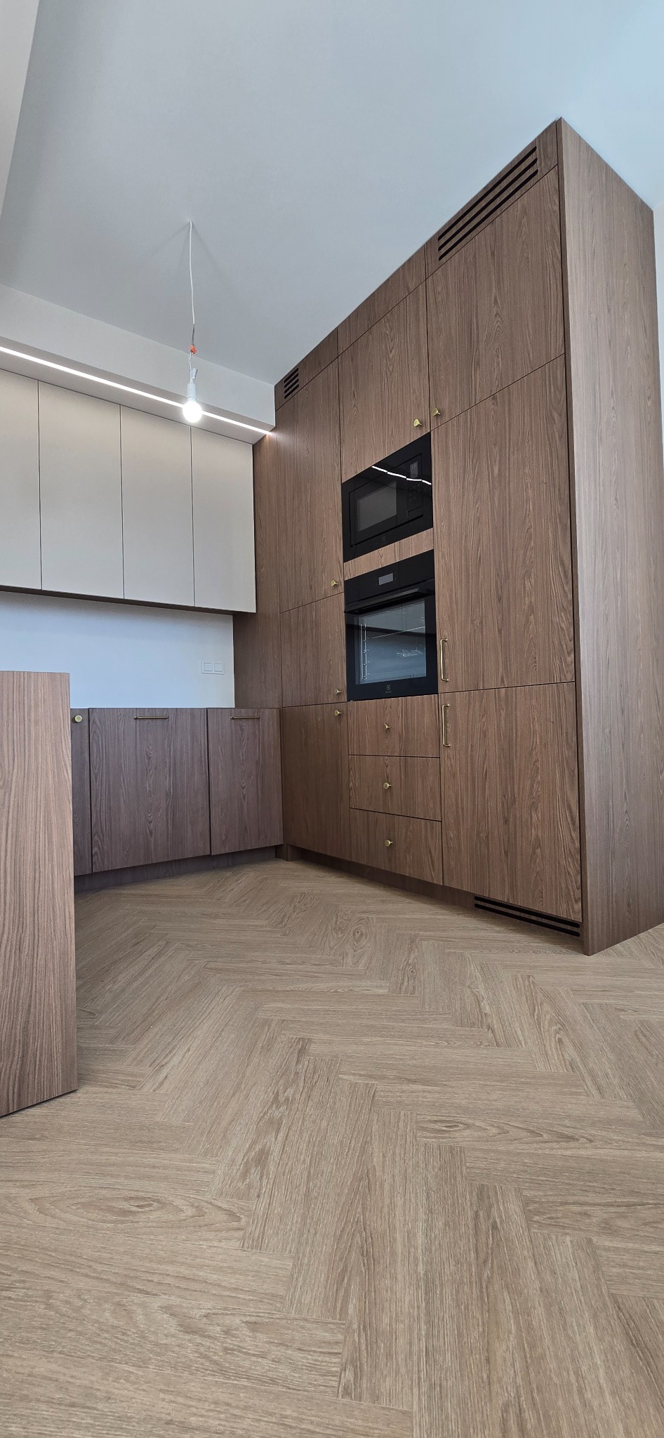 Nowoczesna kuchnia z drewnianymi szafkami, wbudowanymi piekarnikami i mikrofalówką. Parkiet w jodełkę. Minimalistyczne oświetlenie sufitowe.