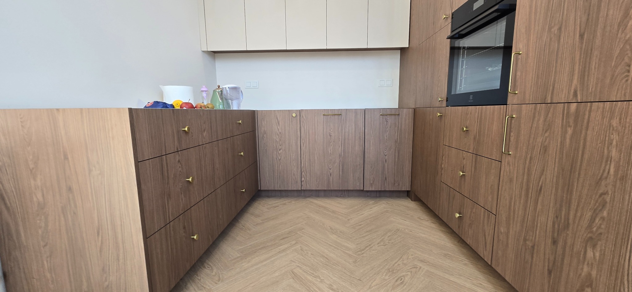 Nowoczesna kuchnia z drewnianymi frontami i złotymi uchwytami, parkiet w jodełkę. Na blacie akcesoria kuchenne i owoce. Minimalistyczny design.