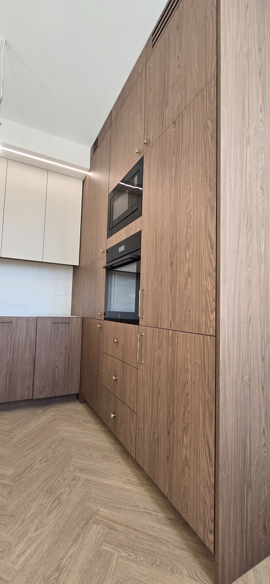 Nowoczesna kuchnia z drewnianymi frontami, złotymi uchwytami i wbudowanym sprzętem AGD. Parkiet ułożony w jodełkę. Ujęcie z perspektywy podłogi.