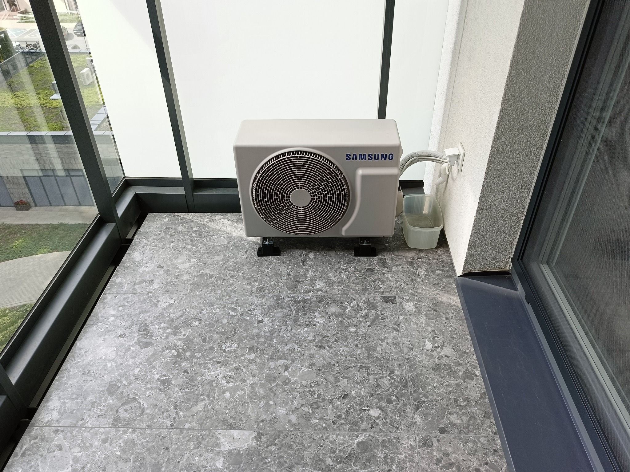 Kompaktowa jednostka zewnętrzna klimatyzacji Samsung na balkonie z szarymi płytkami i widokiem na zieleń za oknem. Widoczne podłączenie do gniazdka.