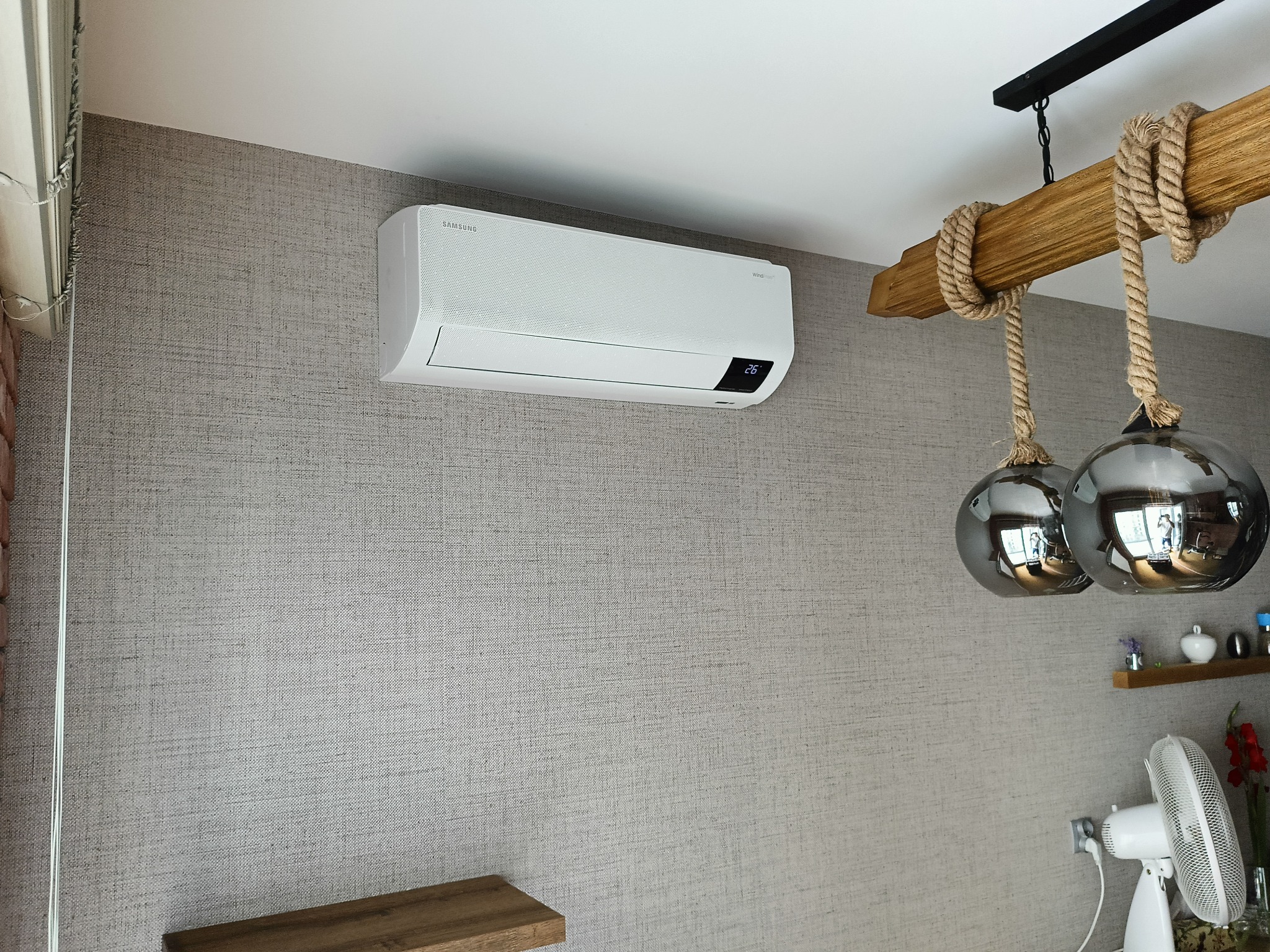 Biały klimatyzator Samsung WindFree zamontowany na szarej ścianie z tapetą o teksturze lnu. W tle lampa z drewnianym elementem i sznurami, wentylator.