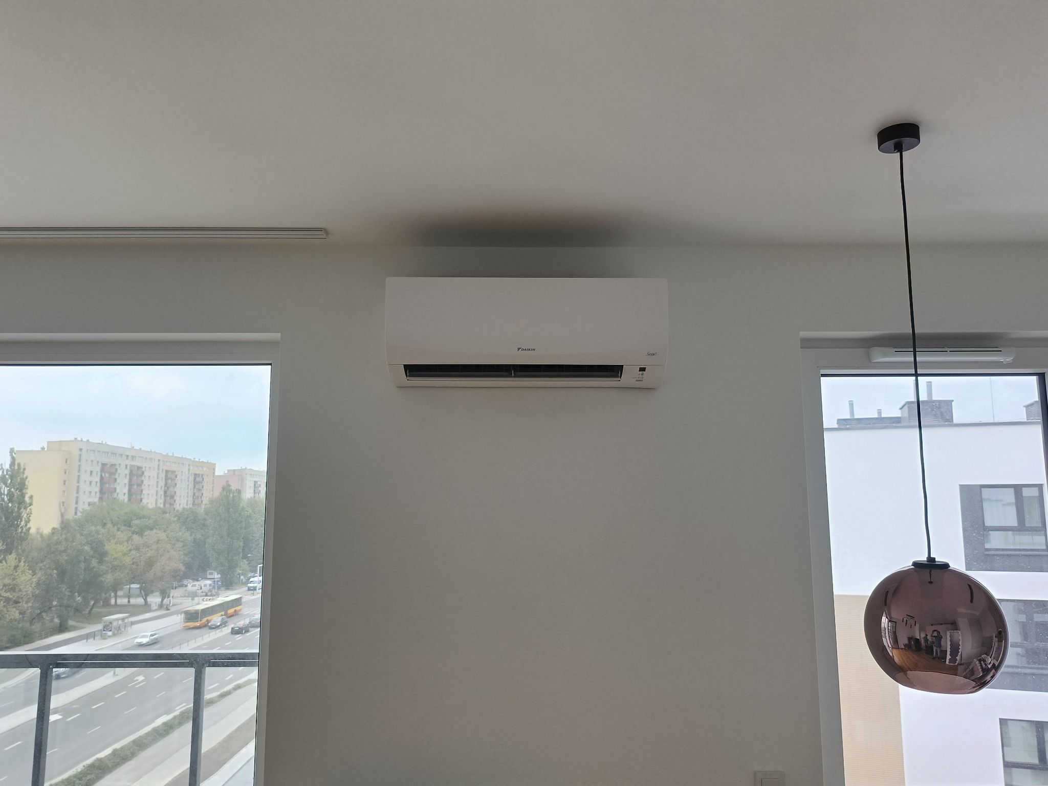 Biała klimatyzacja Daikin na ścianie w jasnym pokoju, widok na miasto przez okno, nowoczesna lampa wisząca.