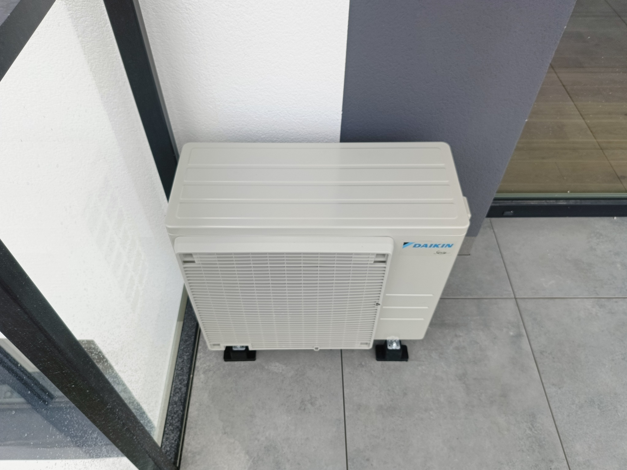 Zewnętrzna jednostka klimatyzacji Daikin zamontowana na szarej podłodze, przy białej ścianie. Widok z góry, ukazujący detale montażu i model klimatyzatora.