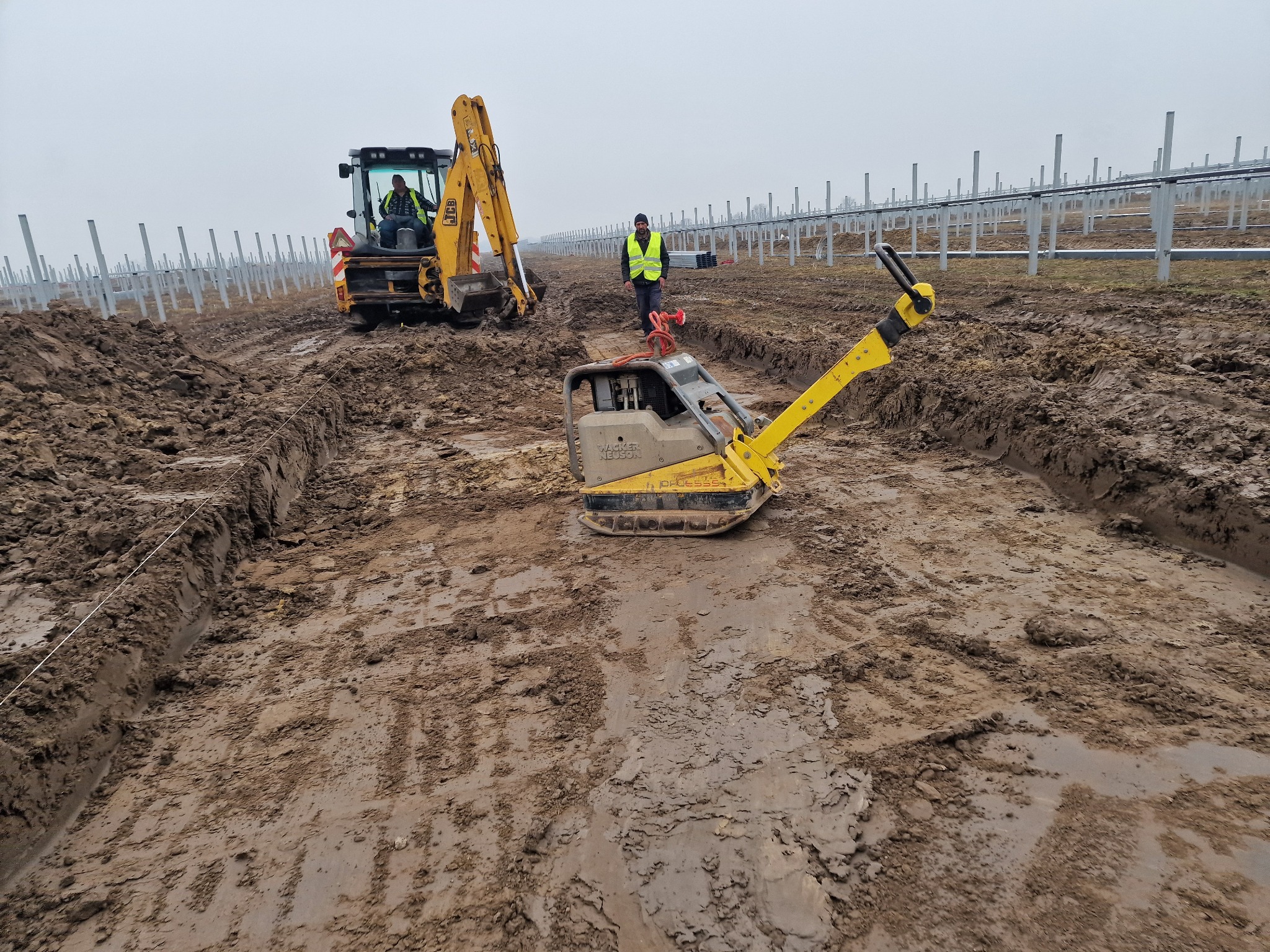 Praca na budowie farmy solarnej: ubijarka, koparka i pracownicy na błotnistym terenie, przygotowującym podłoże pod panele. W tle rzędy słupów konstrukcyjnych.