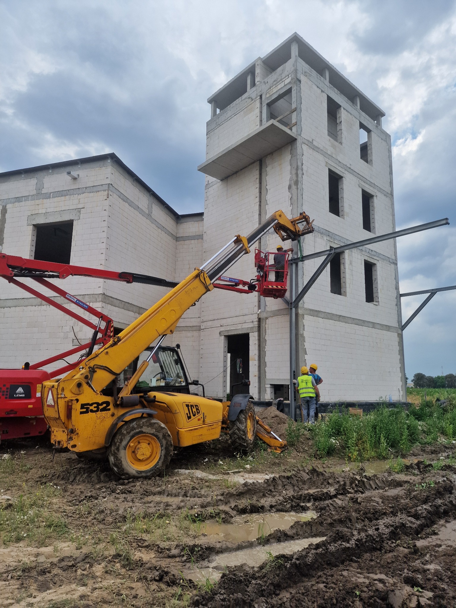 Budowa domu z bloczków: żółta koparka JCB, czerwony podnośnik koszowy i pracownicy montujący stalową konstrukcję na tle budynku w stanie surowym otwartym i pochmurnego nieba.