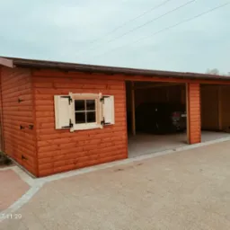 Rustykalna, drewniana konstrukcja z oknem z okiennicami i otwartymi wjazdami, w tle zaparkowany samochód. Widok z zewnątrz, na utwardzonym podłożu.