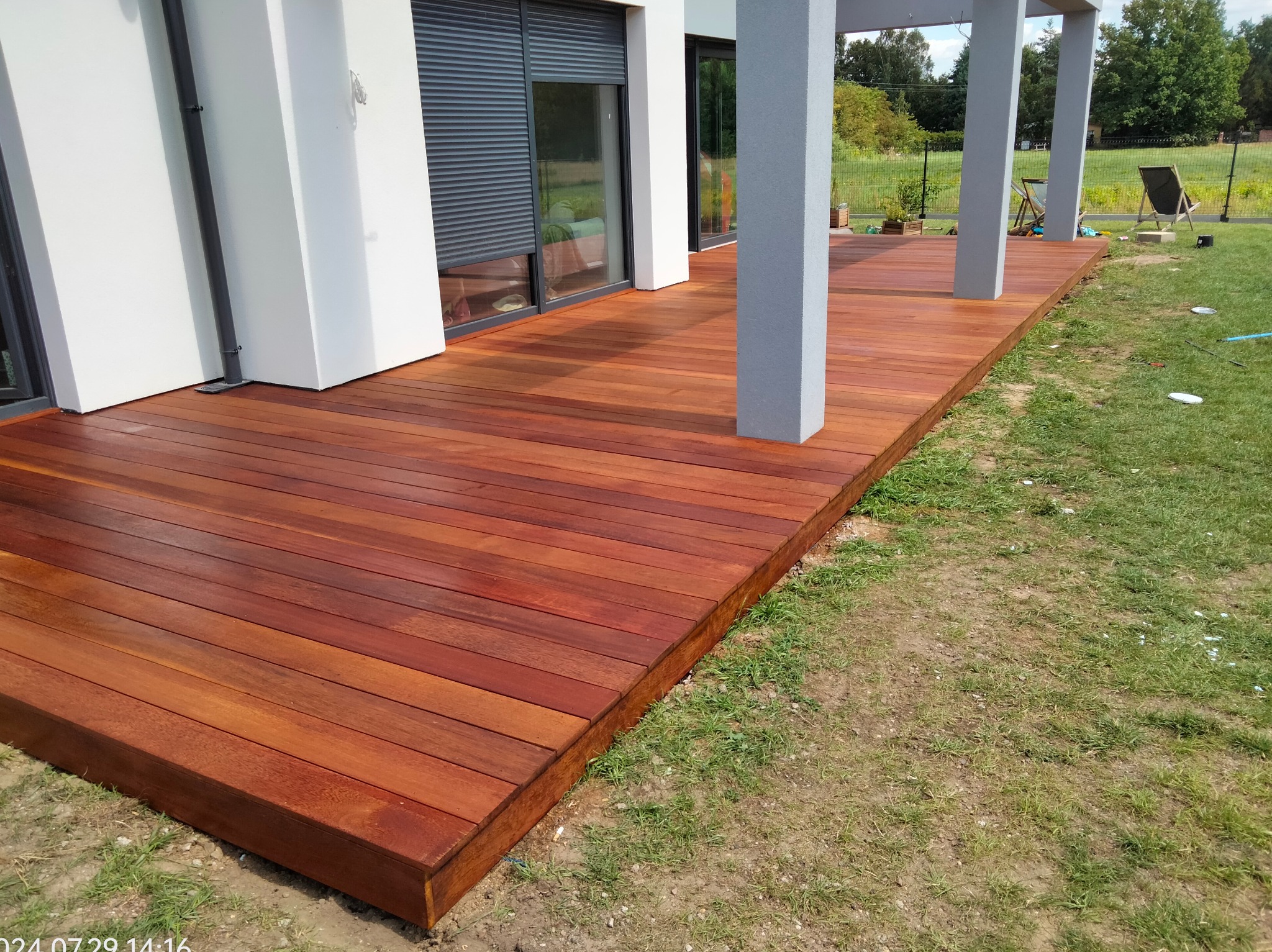 Nowoczesny taras drewniany z egzotycznego drewna, przylegający do białego budynku z szarymi kolumnami, widok na zielony ogród w tle.