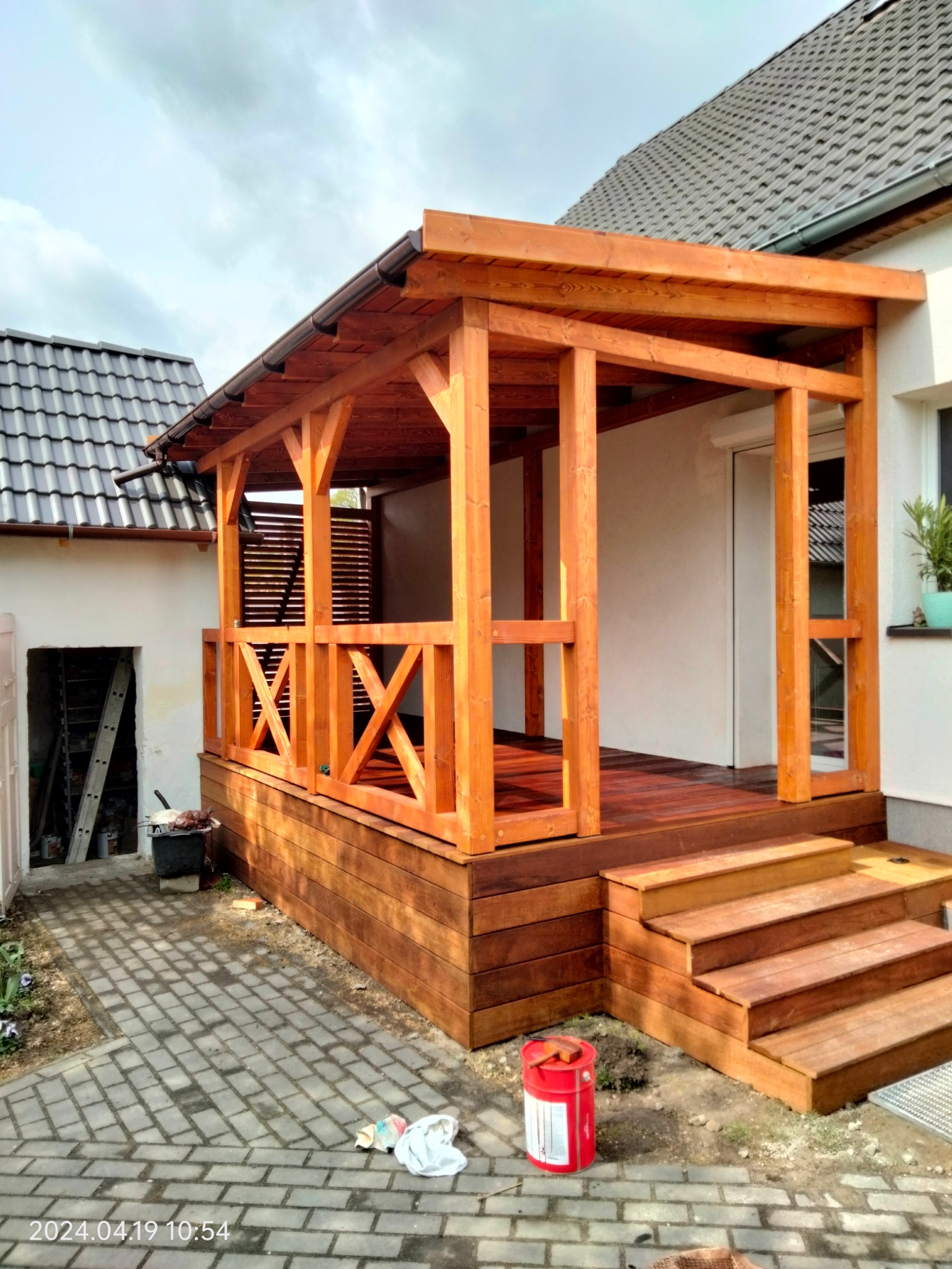 Zadaszony taras z drewna egzotycznego, z balustradą w kształcie litery X. Widoczne schody wejściowe i czerwona puszka z impregnatem. Tło: dom z szarą dachówką.